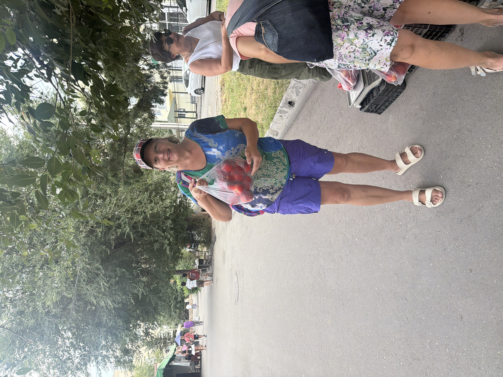 In an outdoor market scene, a woman in a colorful cap and blue-green patterned shirt stands holding a plastic bag filled with red fruit. She wears purple shorts and white sandals. Behind her, another woman in a white tank top and olive green pants, wearing sunglasses, is also visible. In the background, several other people are walking along a paved path lined with trees and market stalls. Cars are parked on a street further in the background, suggesting an urban or suburban location. The ground is a rough asphalt surface, and there is a curb with some vegetation.