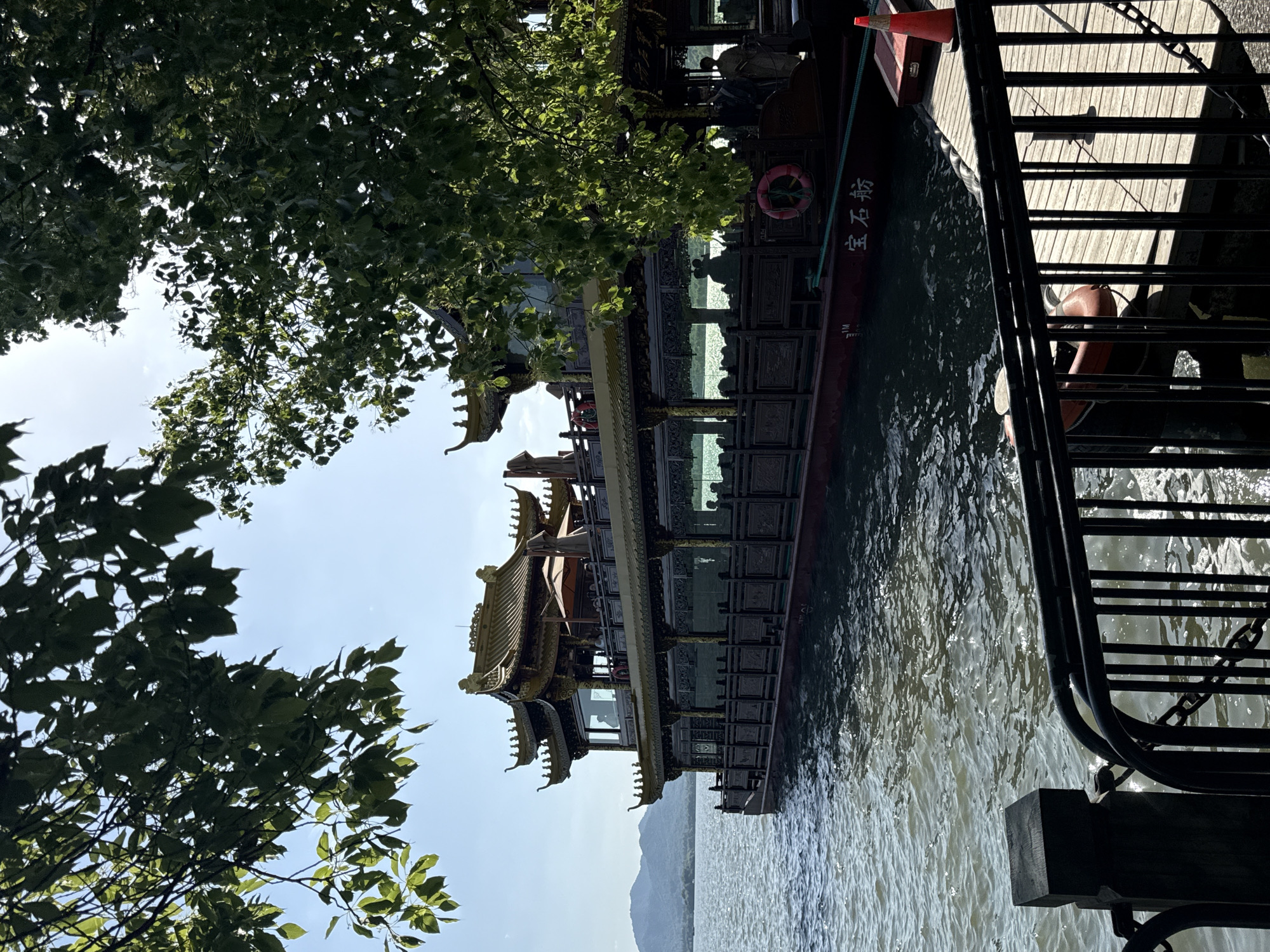 The photograph captures a multi-tiered, ornate boat docked at the West Lake in Shangcheng District, China. The boat features intricate architectural details, including eaves and paneling. A red life preserver is visible on the side of the boat. A metal railing and a wooden dock run along the right side of the frame. Green foliage from nearby trees partially obscures the sky and the boat. In the distance, faint mountains are visible across the lake. The water surface has ripples and light reflections.