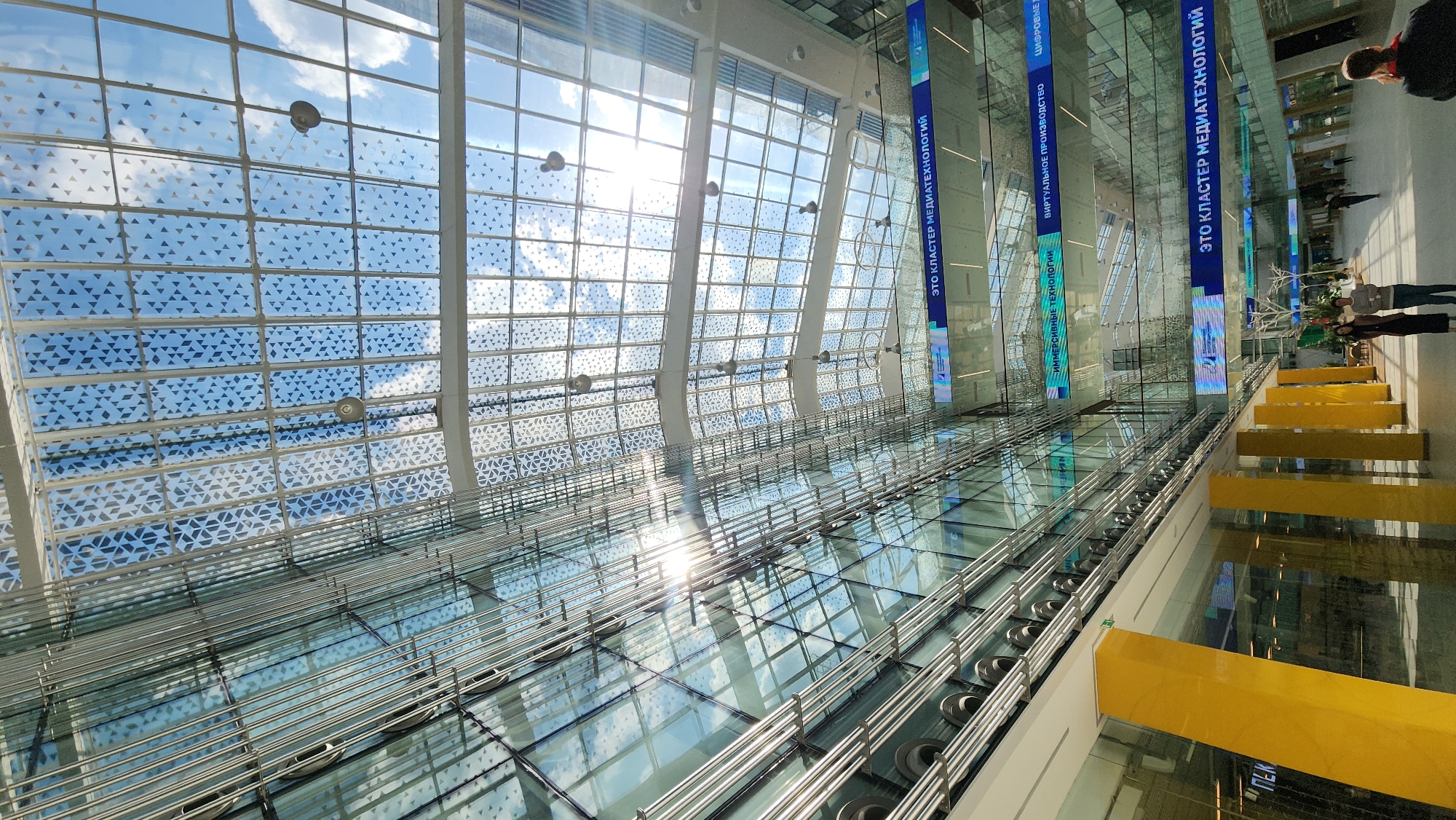The image depicts the interior of a building in Moscow, Russia. A large glass ceiling with a repeating triangular pattern allows sunlight to stream into the building. Overhead, several blue banners with the text "ЭТО КЛАСТЕР МЕДИАТЕХНОЛОГИЙ" (This is the Media Technology Cluster) hang suspended. Underneath the glass ceiling, a glass walkway is supported by a metal framework. A few people are visible on the right side of the image. A series of vertical yellow bars are spaced along the right side of the walkway. The image is taken from a low angle, looking upwards towards the ceiling.