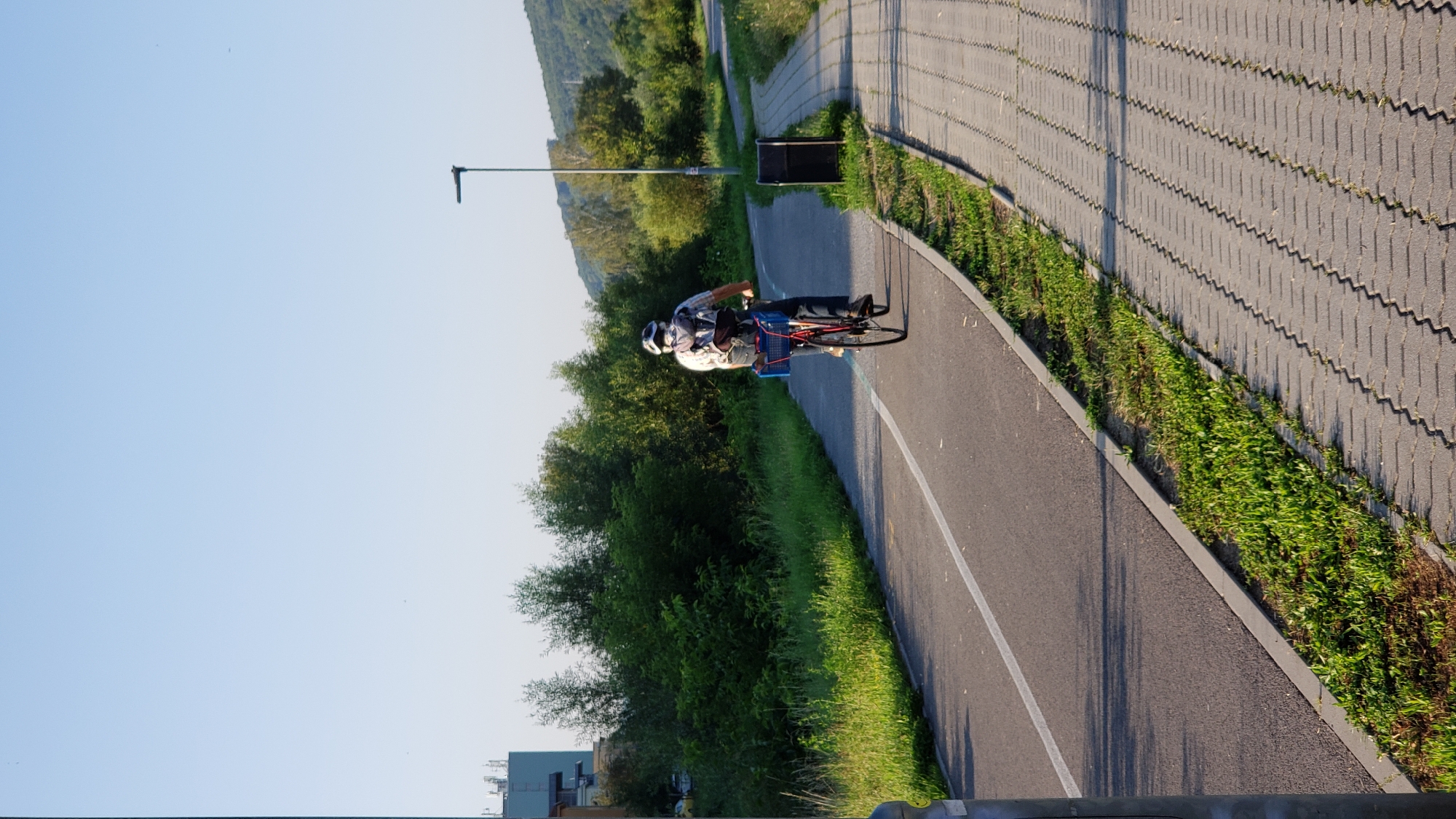 A person wearing a helmet and a backpack is riding a bicycle on a paved bike path. The bicycle has a blue basket attached to the back. The path is lined with grass and trees, with a paved sidewalk on the right. In the background, there are buildings and a hill. The scene appears to be in a park or recreational area. This scene is located on the bike path near the bus station in Prešov, Slovakia.