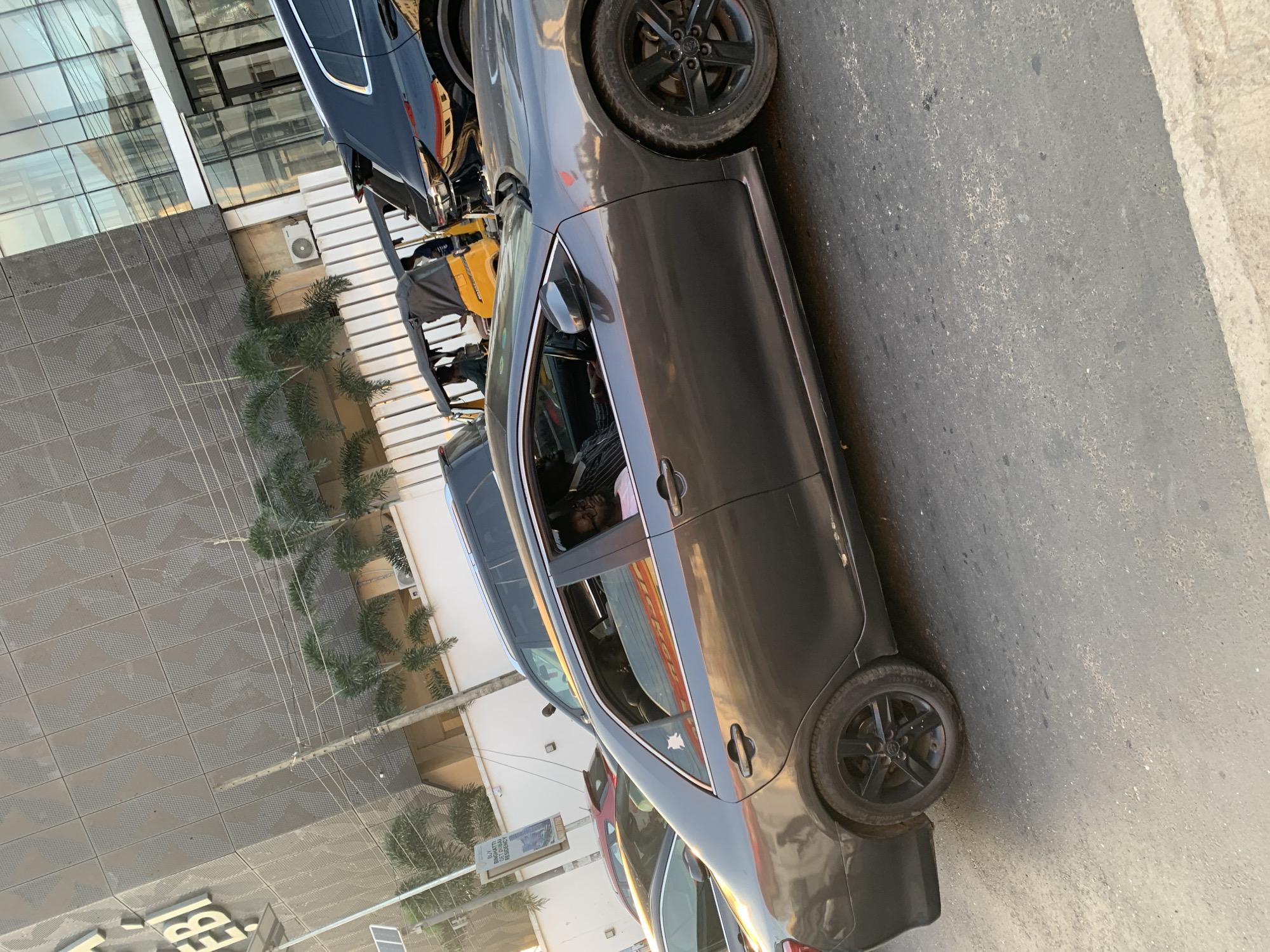 The image captures a street scene in Ikeja, Nigeria, with several vehicles and a modern building in the background. In the foreground, a dark grey sedan is prominently featured, with its driver visible through the open window. The driver is a Black male wearing glasses and a light-colored shirt. The car has black rims on its wheels. Beside the sedan, to its right, is a black SUV. To the left of the sedan, other vehicles are partially visible, including a red car and another sedan. In the middle ground, a yellow auto-rickshaw, also known as a "Keke Napep," is present, with passengers inside and a person driving it. Behind the vehicles, a building with a modern facade, featuring patterned metal panels and glass windows, stands tall. Palm trees are scattered in front of the building. Overhead, power lines crisscross the scene. The overall impression is of a busy urban environment, likely during daylight hours, with traffic on a paved road.