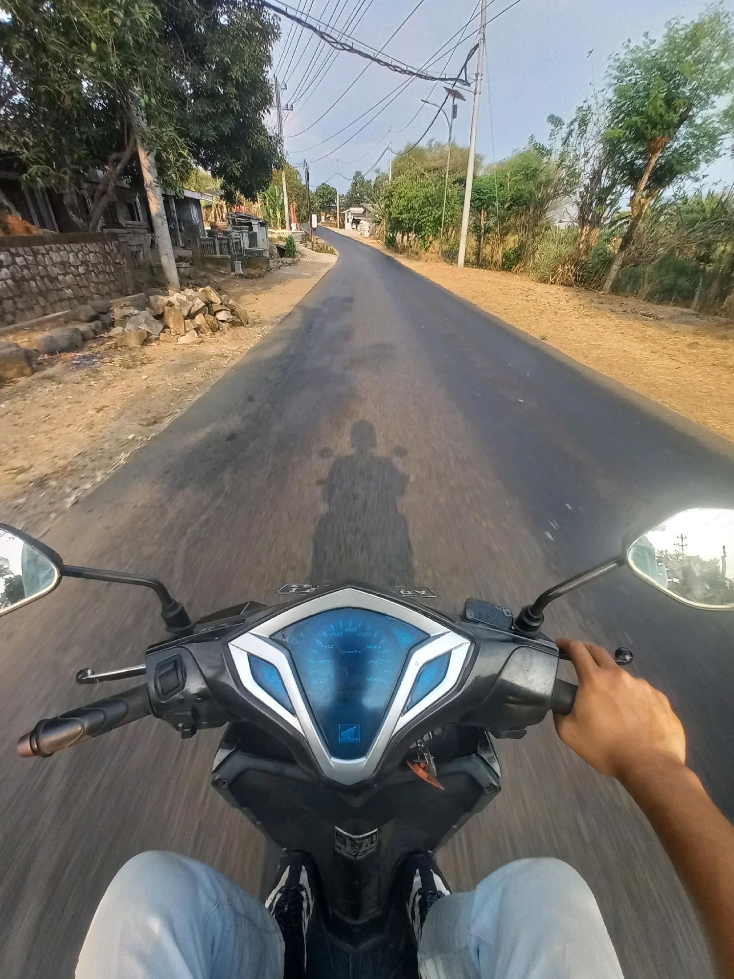 A first-person perspective shot captures a person riding a motorcycle down a paved road in Kulurejo, Indonesia. The rider's hands grip the handlebars, with one hand resting on the throttle. The motorcycle's dashboard, displaying speed in km/h, is visible in the foreground. The road is flanked by a dry, dusty embankment on the left and a stretch of trees and vegetation on the right. In the distance, electrical poles and wires crisscross the sky. The scene appears to be a rural or semi-rural area, possibly a village or outskirts of a town, given the presence of simple houses and scattered trees. The lighting suggests it is either early morning or late afternoon, with the sun casting shadows on the road.