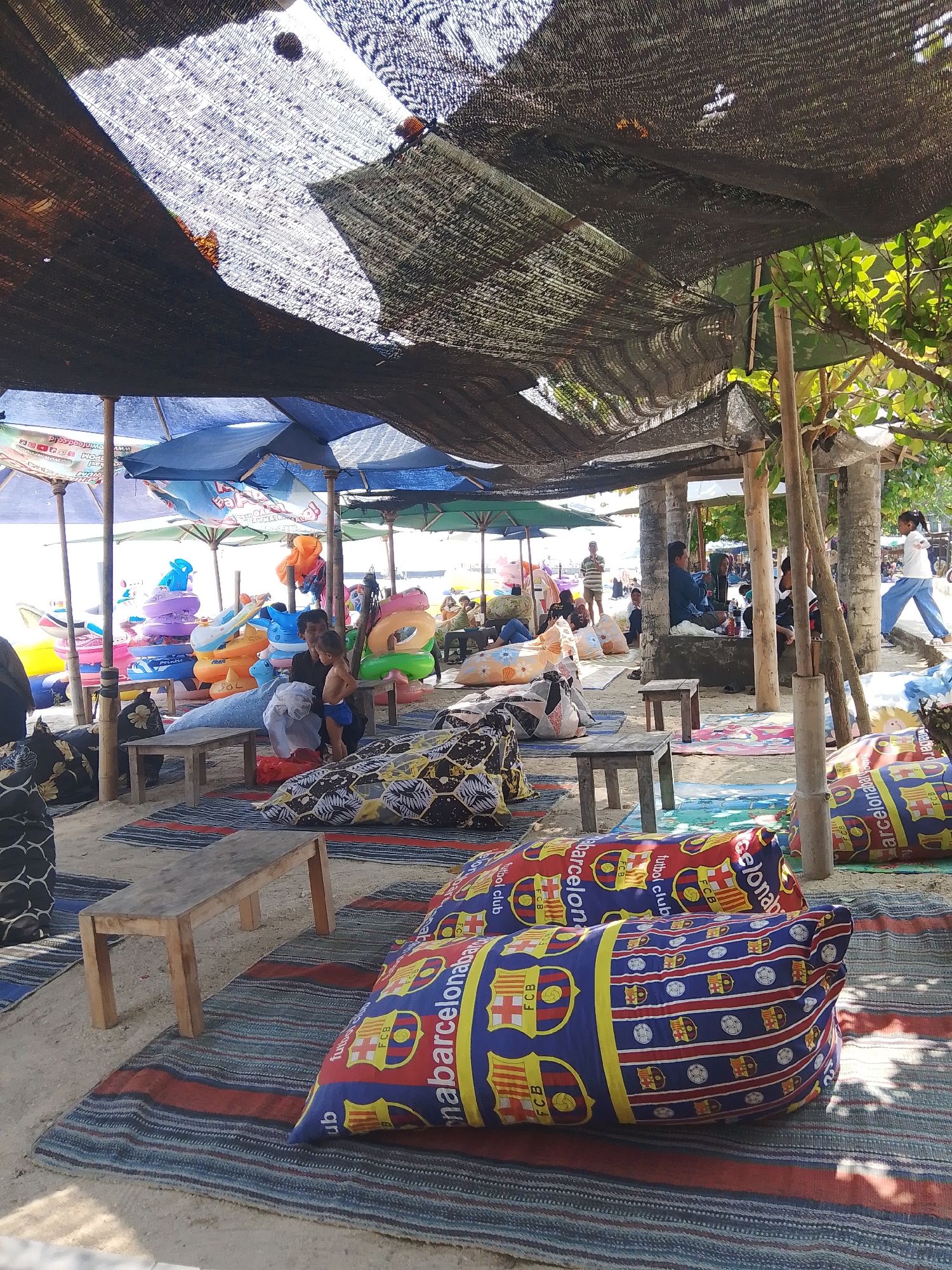 The image depicts a beachfront seating area at the beach in Pejagan, Indonesia. Several rows of colorful beanbag chairs are arranged on striped mats spread across the sandy ground. In the background, a collection of inflatable rings in various sizes and colors are stacked. A man holds a young child, who is partially visible, near a concrete structure. Other individuals are seated or standing in shaded areas created by awnings and beach umbrellas. A woman in white and blue clothing walks across the sand. The scene is illuminated by natural sunlight filtering through the overhead coverings.