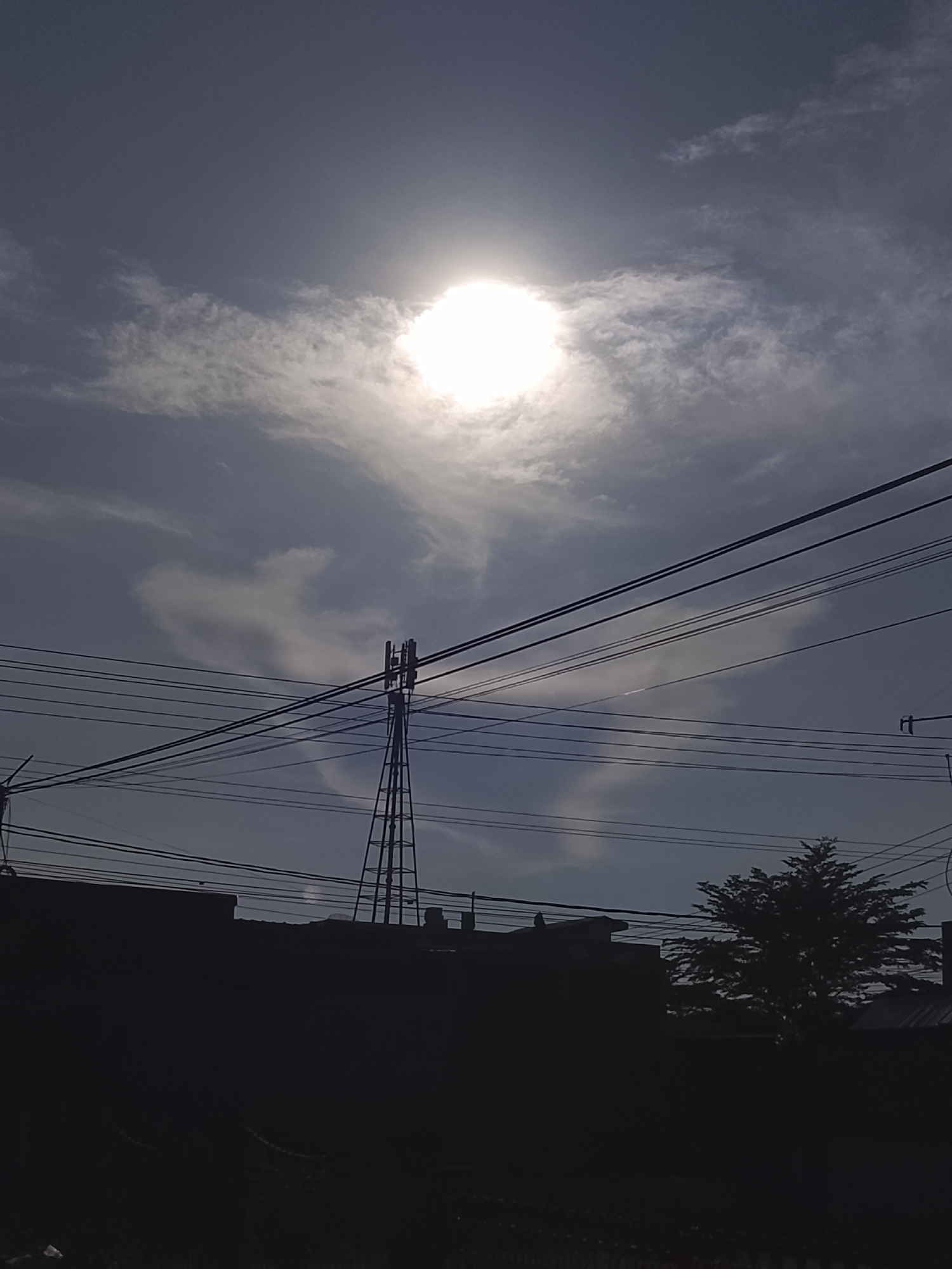 A transmission tower stands silhouetted against a bright, hazy sun and a cloudy sky. Several power lines crisscross the frame in front of the tower and the sky. In the foreground, the dark silhouettes of buildings and a tree can be seen. The scene is likely located in Lhokseumawe, Indonesia, given the presence of transmission towers and the general appearance of the buildings.