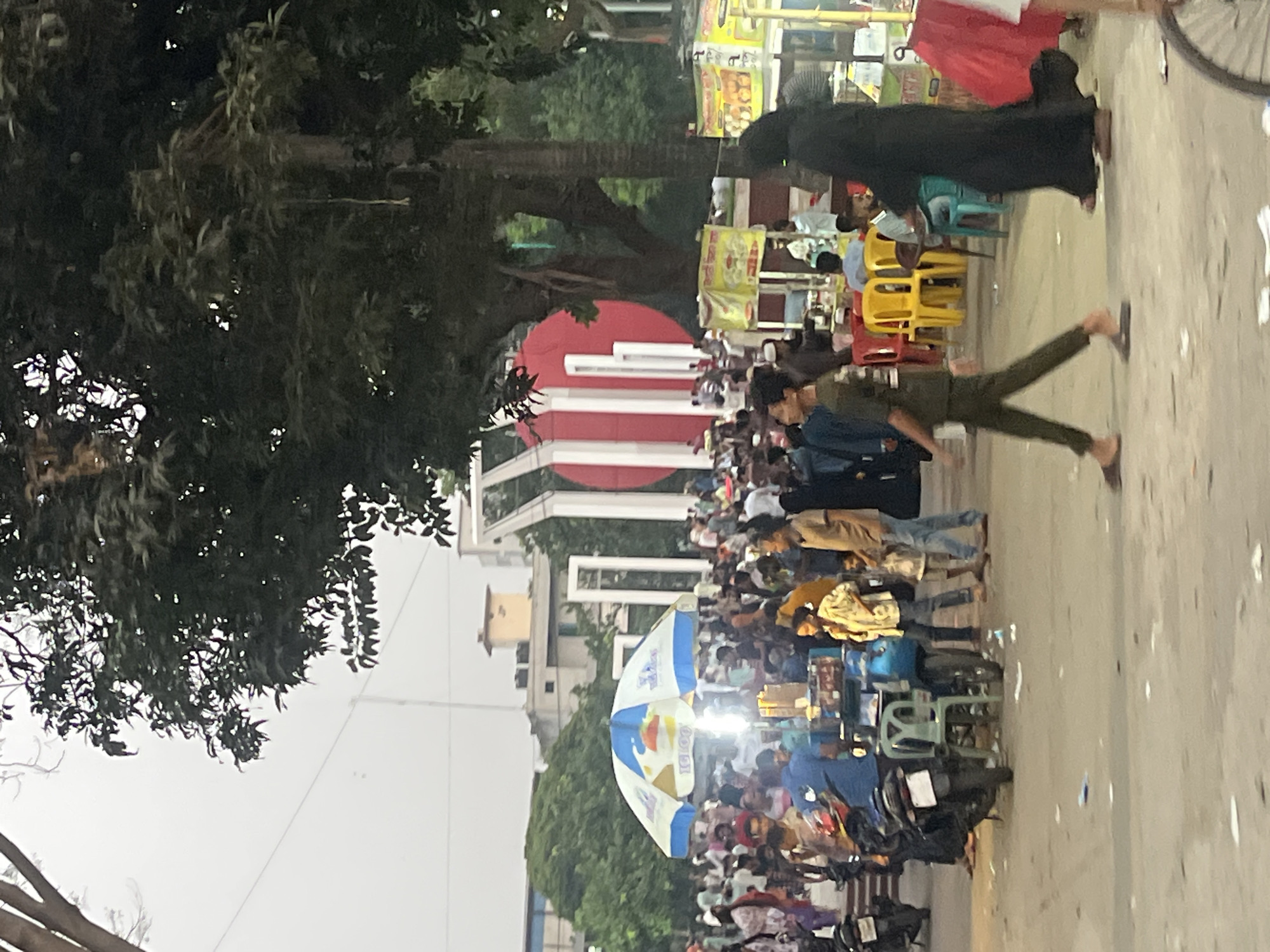 The image depicts a bustling street scene in Dhaka, Bangladesh. The scene is crowded with people, some walking, some seated at food stalls, and others on motorbikes. Many food stalls are visible, with yellow chairs and colorful signs. A large tree dominates the left side of the frame. In the background, there's a building with a red and white circular design. The sky is overcast.