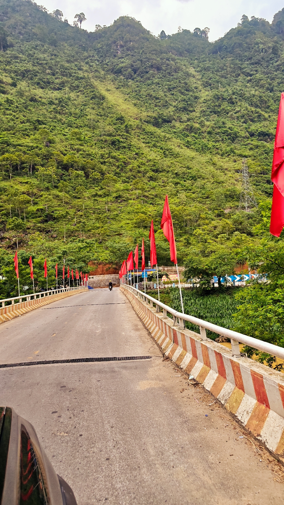 The image shows a bridge in Cán Tỷ Commune, Vietnam, decorated with red flags. A road stretches across the bridge, with a motorcyclist in the distance. The bridge has a white railing and a barrier with red and tan diagonal stripes. A steep, green, tree-covered mountain forms the backdrop. Power lines are visible on the mountainside. Vegetation is present along the sides of the bridge. A portion of a vehicle is visible in the foreground. The sky appears overcast.