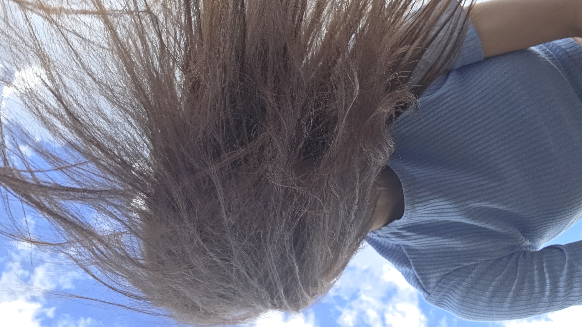 A person's head and shoulders are visible from a low-angle perspective, with their long, light brown hair blurred and blowing in the wind across the frame. The person is wearing a light blue, ribbed, long-sleeved shirt. The background consists of a bright blue sky with scattered white clouds. There is no discernible city or specific location information visible in the image.