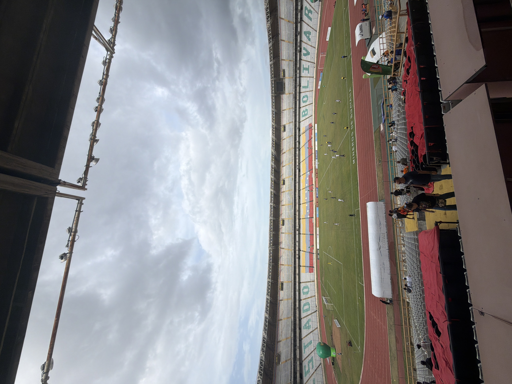 The image shows a soccer game in progress at the CTE Cachamay stadium in Ciudad Guayana, Venezuela. The stadium has tiered seating, some of which is covered with red fabric. The word "BOLÍVAR" is visible on the stadium seating. There are people watching the game from the stands, and the weather appears overcast.