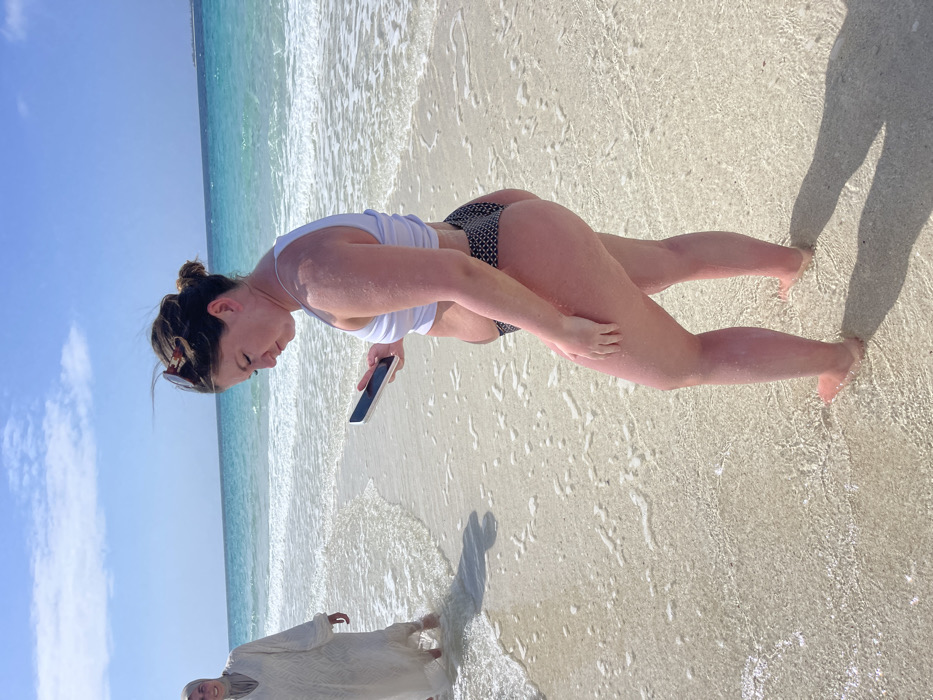 The image depicts a bright, sunlit beach scene in Zanzibar, Tanzania. In the foreground, a woman stands facing left in profile, barefoot in shallow, clear ocean water. She wears a white sleeveless crop top and dark-patterned bikini bottoms. Her brown hair is pulled back, with sunglasses resting on her head. She holds a white smartphone with both hands, looking down at its screen. Her skin, particularly on her legs, exhibits a reddish tone consistent with sun exposure.
In the mid-ground, to the left, another individual stands partially submerged in the shallow water. This person wears a light-colored, long-sleeved abaya or dress with a matching head covering (hijab) and is smiling while looking towards the foreground woman.
The environment features a white sandy beach with gentle waves lapping at the shore. The ocean water transitions from clear turquoise near the beach to a deeper blue further out. The sky above is clear blue with minimal white clouds. Strong daylight casts distinct shadows from the figures onto the wet sand. A faint landmass or structure is visible on the distant horizon to the right.