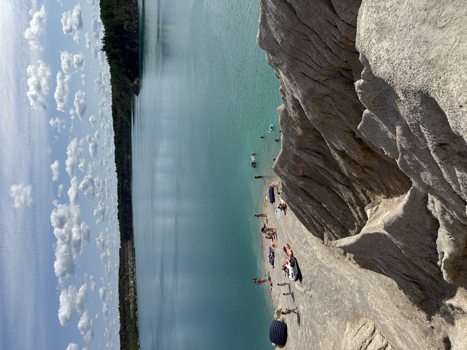 An elevated long shot captures a scene at a quarry lake in Druzhba, Ukraine, under a partly cloudy sky. The foreground consists of steeply eroded, light beige to grey rock and sand formations with sharp, jagged ridges. Below these formations, a narrow, light-colored sandy beach extends along the edge of a large body of highly saturated turquoise water.
Approximately 20-30 individuals are present on the beach and in the water. On the beach, people are seen standing, sitting, and lying on towels. Notable objects include a large dark blue inflatable mattress, several smaller inflatable mats, and a small inflatable boat with an attached motor. In the water, individuals are swimming or wading, with some utilizing inflatable rings and floats.
The midground features the expansive, calm turquoise water. The background reveals a distant shoreline densely covered with dark green trees, with some lighter rock or earth exposures visible along the banks. The sky above is bright blue with scattered white cumulus clouds.
