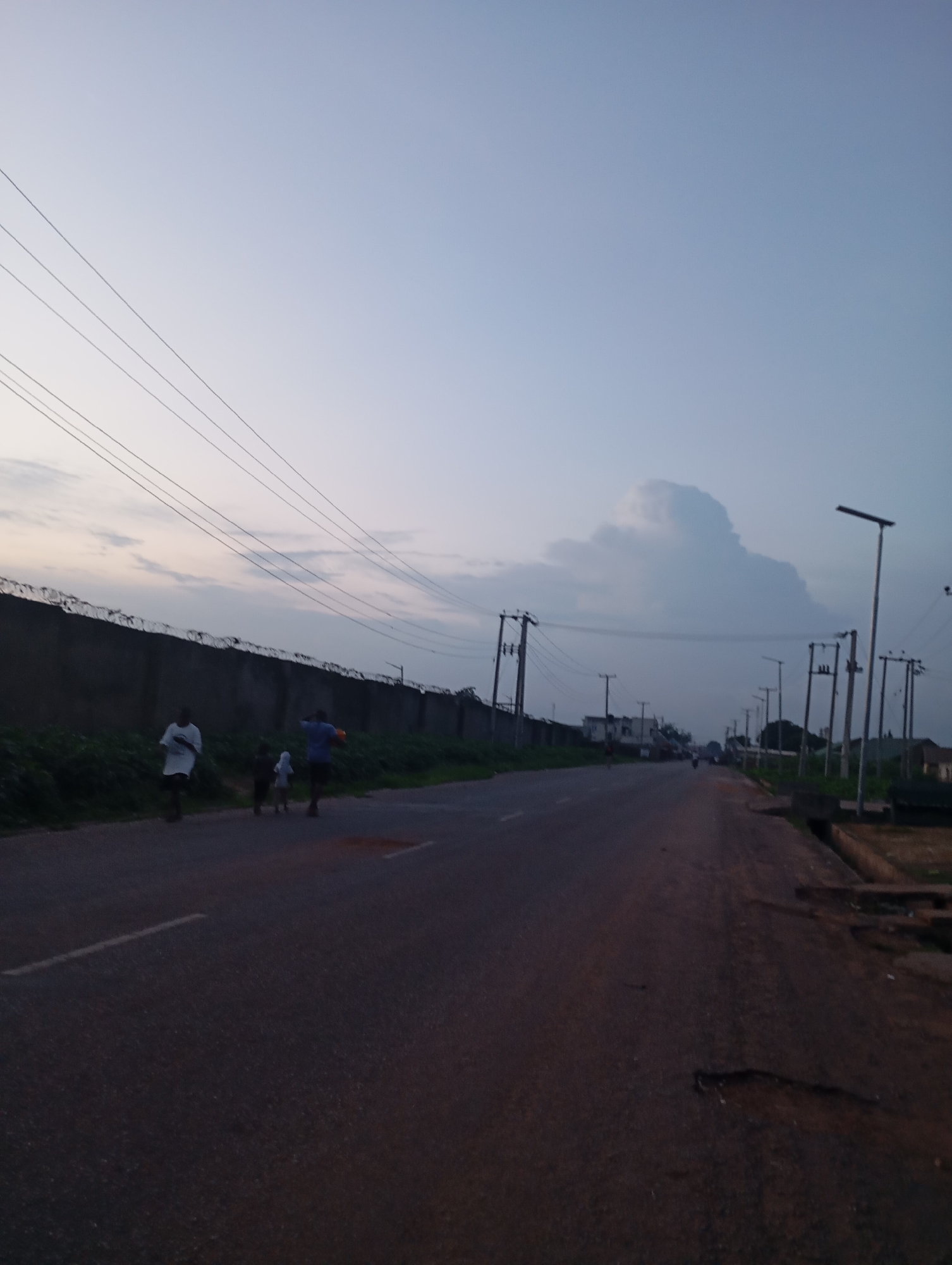 A group of people walk along a paved road in Shendam, Nigeria, during twilight. The road is lined by a grey concrete wall topped with barbed wire on the left, and sparse vegetation. Several utility poles with wires are visible along the road. In the distance, a large, puffy cloud is visible in the pale sky, along with more utility poles and distant buildings. The road surface is dark and shows signs of wear and tear, with some patches of red dirt. The group of people includes two adults and two children, with one adult carrying an orange object that appears to be a ball. They are walking away from the camera, towards the distant part of the road.
