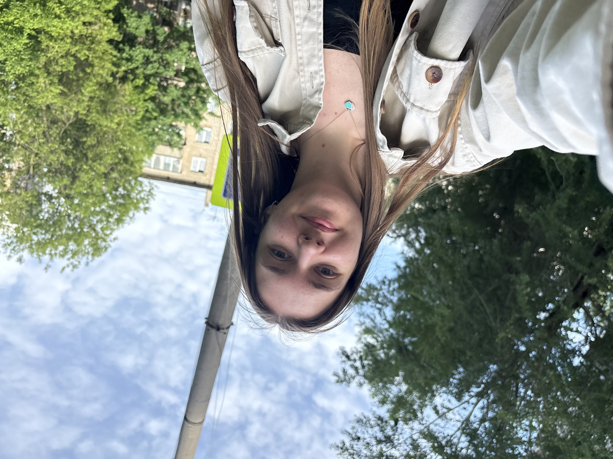 A photo taken outdoors, showing a young woman with long brown hair wearing a beige jacket and a necklace with a turquoise pendant. She is standing near a gray concrete pole with a yellow and black sign attached. The background shows green trees, a building with windows, and a sky with scattered white clouds. The location is Perm, Russia.