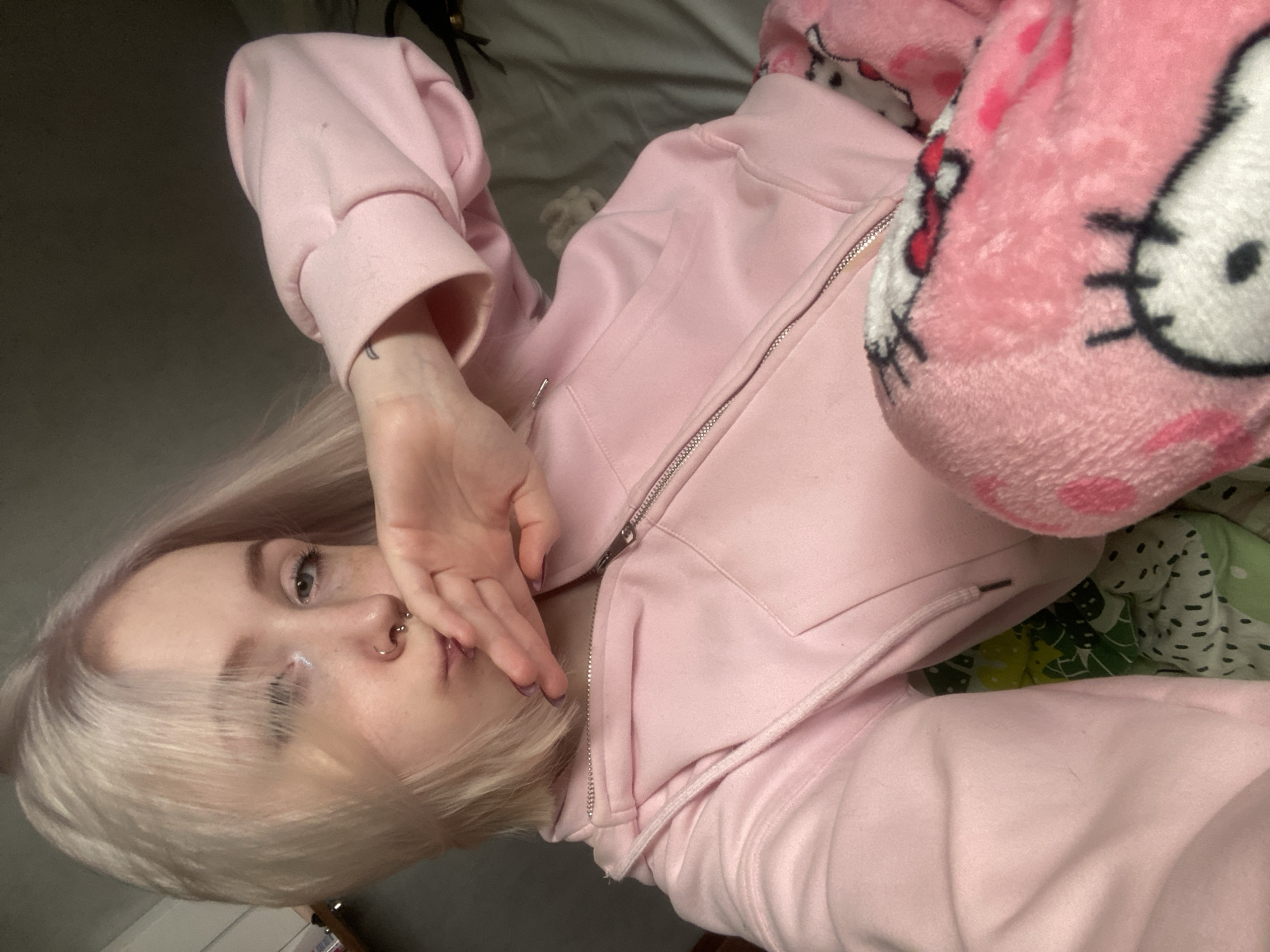 A fair-skinned individual with pale blonde hair is depicted lying on their back in an indoor setting, likely taking a selfie from an overhead angle. The subject, who appears to be young and female, is wearing a light pink zippered hoodie and fuzzy pink sleepwear or pants adorned with large Hello Kitty faces. One hand is raised, partially covering their mouth and chin.
Notable features of the individual include light-colored eyes, a septum piercing, and pink/purple nail polish visible on their fingers. A small, dark tattoo is visible on their right wrist. The background is muted, showing hints of gray and a green patterned fabric, suggesting a bed or couch. The overall aesthetic is soft and relaxed. No specific time of day or weather can be discerned, but the indoor setting implies artificial lighting or indirect natural light. There is no visible text in the image. The location context provided is Izhevsk, Russia.