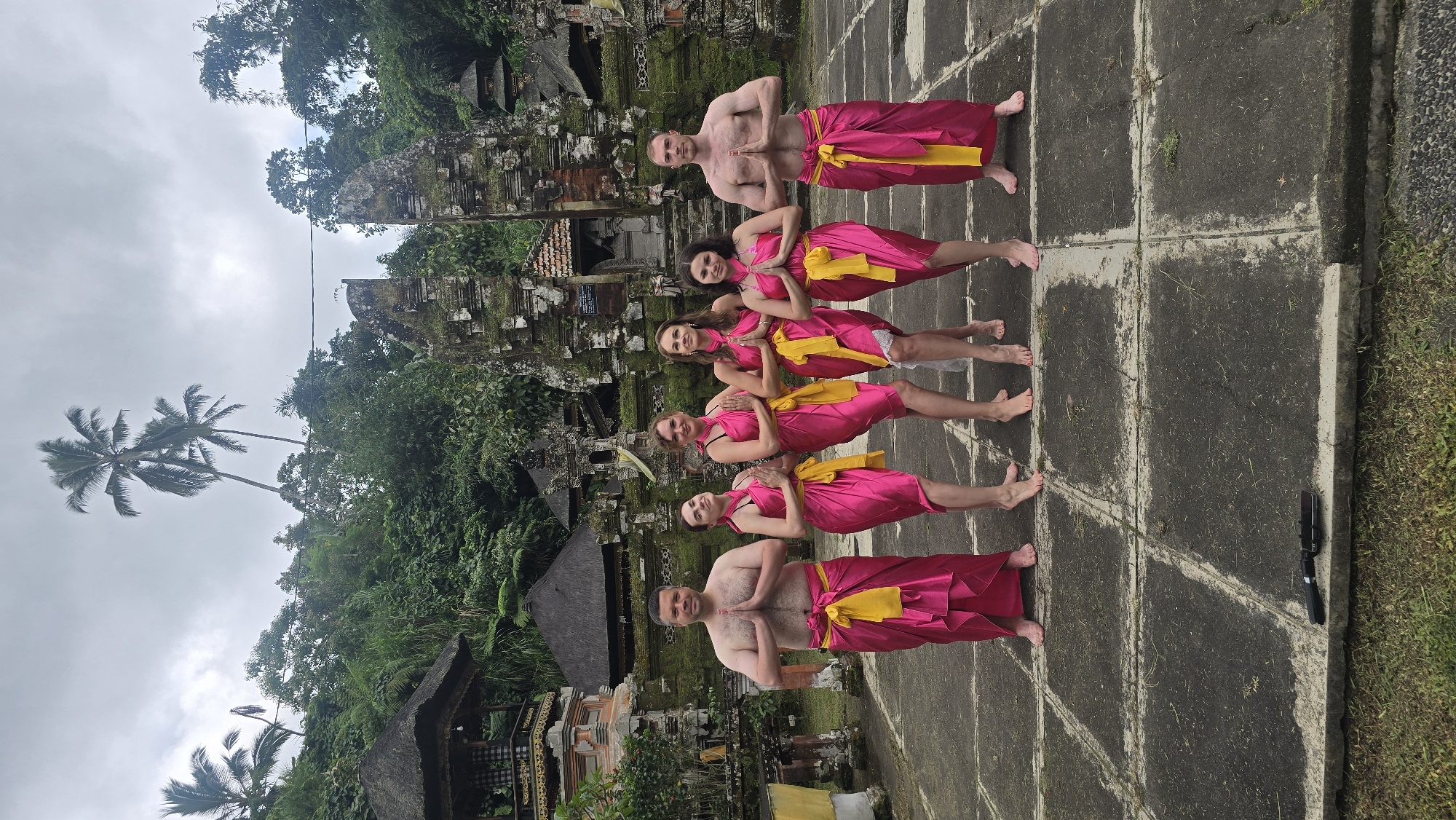 A medium shot captures a group of five individuals standing barefoot in a line, facing the camera with hands pressed together in front of their chests. They are clad in pink sarongs secured with yellow sashes, against the backdrop of a traditional Balinese temple. The temple, constructed from weathered stone, is adorned with lush vegetation.
The scene is set on a stone pavement with visible grid-like patterns. To the left of the frame, there is a cluster of palm trees and greenery under a cloudy sky, which suggests it might be overcast.
Based on the architectural style of the temple, the tropical flora, and traditional clothing, the location is likely in or around Tegallalang, Indonesia. A black phone stand can be seen in the bottom right corner.
