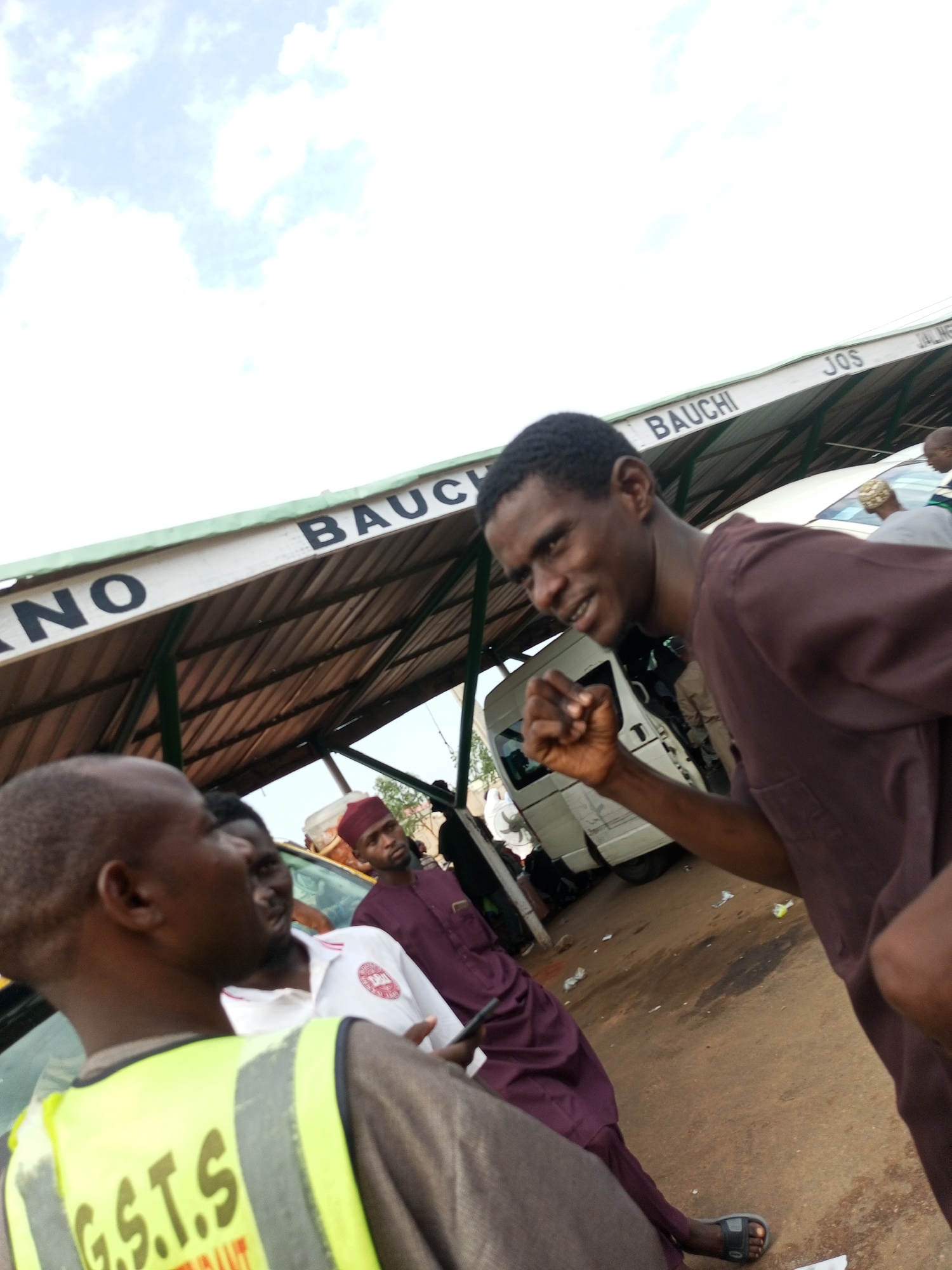 In Gombe, Nigeria, a group of people are gathered under a covered structure, likely a bus station or market shelter. The structure has a metal roof supported by green metal beams and a white fascia board with black lettering. The words "BAUCHI" and "JOS" are visible on the fascia, suggesting these are common destinations or routes from this location.
In the foreground, a man wearing a bright yellow high-visibility vest over a brown shirt is partially visible from the back and side, facing away from the camera. The vest has the inscription "G.S.T.S." in large green letters.
To the right, a man in a brown tunic is in focus, facing the camera with a determined expression and a clenched fist raised slightly. His right arm is bent at the elbow, and his hand is partially open.
Behind him, a man wearing a dark red cap and a matching maroon garment stands looking forward.
Further back, several other individuals are present, some partially obscured by the foreground subjects and the structure. A white van is visible to the left of the man in the brown tunic, its doors open. The ground is dusty and appears to be a dirt or gravel surface, with some scattered debris. The sky above is bright and partly cloudy. The overall scene suggests a busy transportation hub or public gathering space within the city of Gombe.
