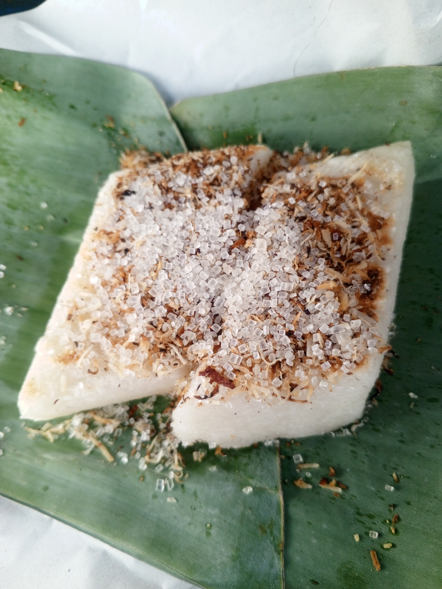 A close-up, top-down shot shows a portion of a flat, white confection, possibly rice cake, resting on a large, vibrant green banana leaf. The confection is topped with a generous scattering of coarse white sugar crystals and toasted shredded coconut, which is brown and granular. Some of the sugar and coconut mixture has fallen onto the banana leaf surrounding the confection. The background is blurred and appears to be white paper. The overall scene is likely from a street food stall or a home kitchen in Depok, Indonesia, where such traditional sweets are commonly prepared and served.