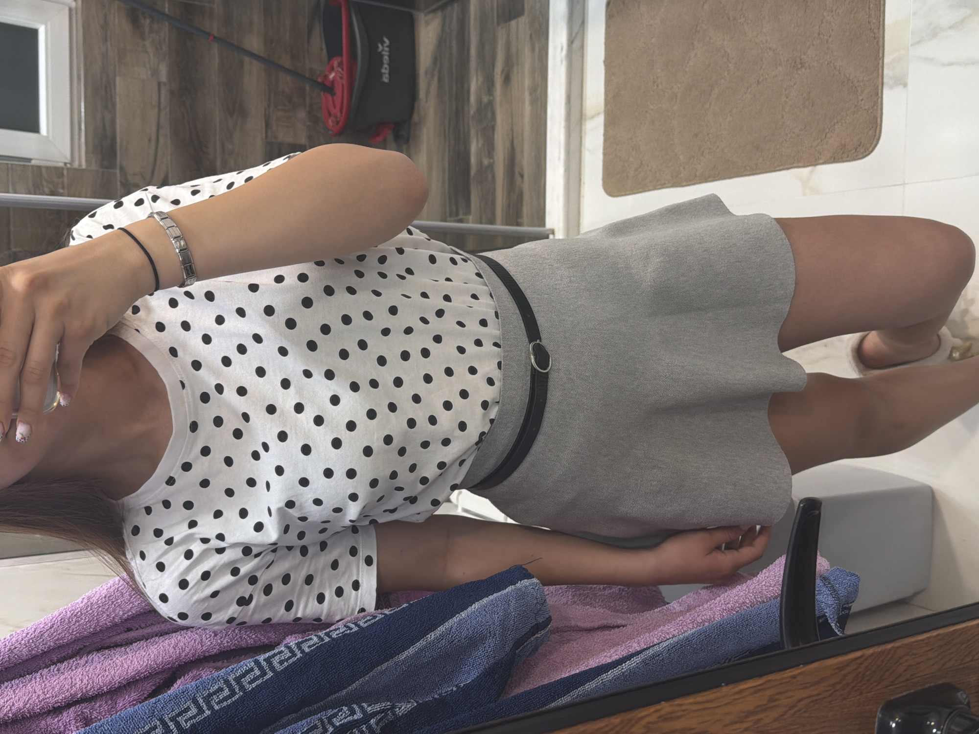 A person is visible indoors. The person is wearing a white polka-dot top, a gray skirt and a black belt. On their left wrist they are wearing two bracelets, a chain bracelet and a band bracelet. The person is on a surface covered with a purple and blue towel. Behind the person there is a beige bath mat, a window, a gray wall, and a red and black object attached to the wall. The city of Buhovo, Bulgaria is not visibly apparent.