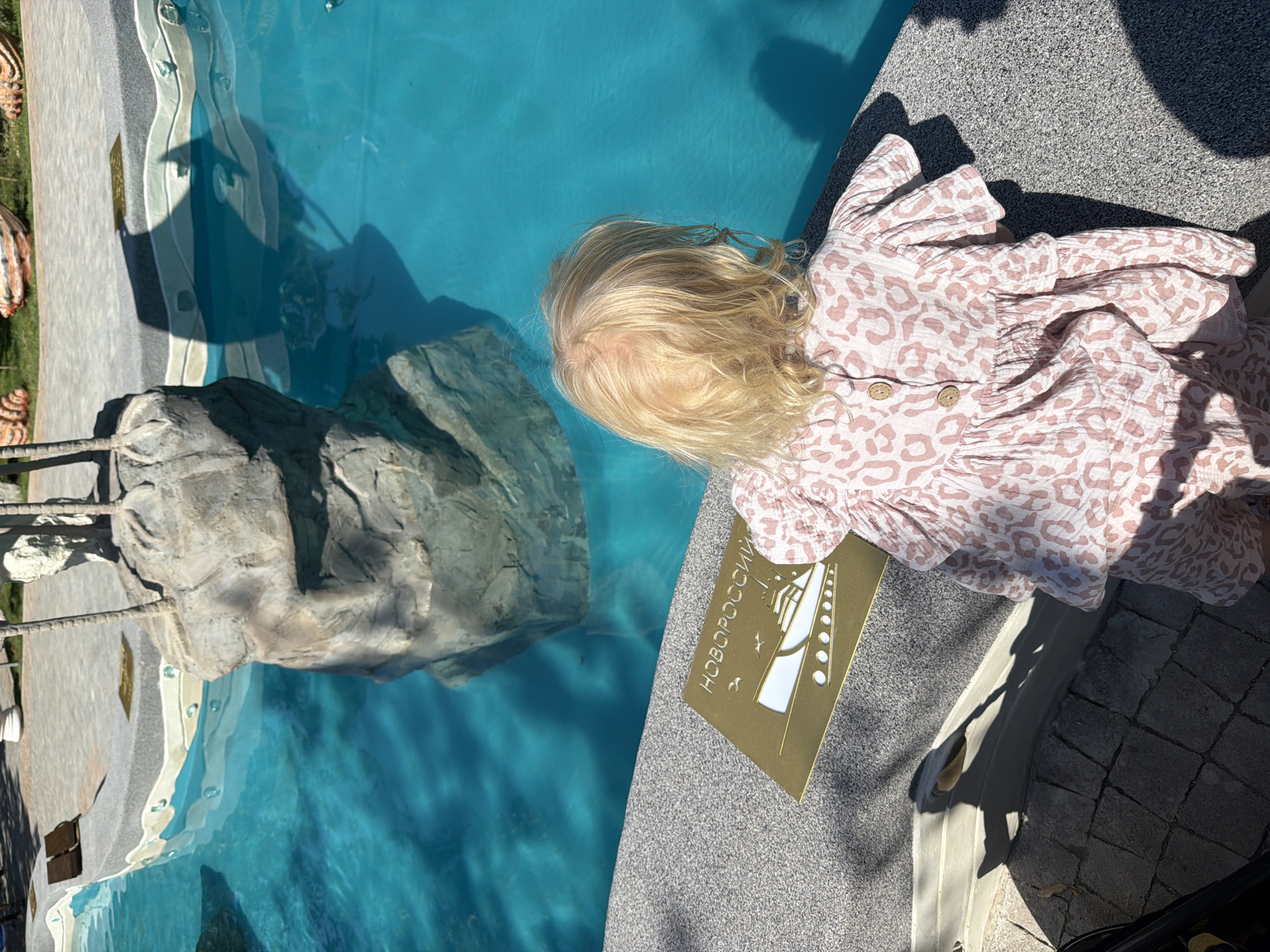 A young Caucasian child with blonde hair, seen from behind, wearing a light pink leopard-print romper, stands at the edge of a vibrant turquoise pool. The child is looking at a golden plaque placed on the grey stone border of the pool. The plaque features a silhouette of a ship and the Cyrillic text "НОВОРОССИЙ". In the background, artificial rocks and palm trees are visible. The scene appears to be outdoors on a sunny day, with shadows cast by the rocks and the child onto the stone and water. This is likely an attraction or a decorative element within a public space. Given the text on the plaque, the location is likely related to Novorossiysk, a city in Krasnodar Krai, Russia. The presence of a pool with artificial rock formations suggests it could be a water park, a resort, or a themed area in a park.