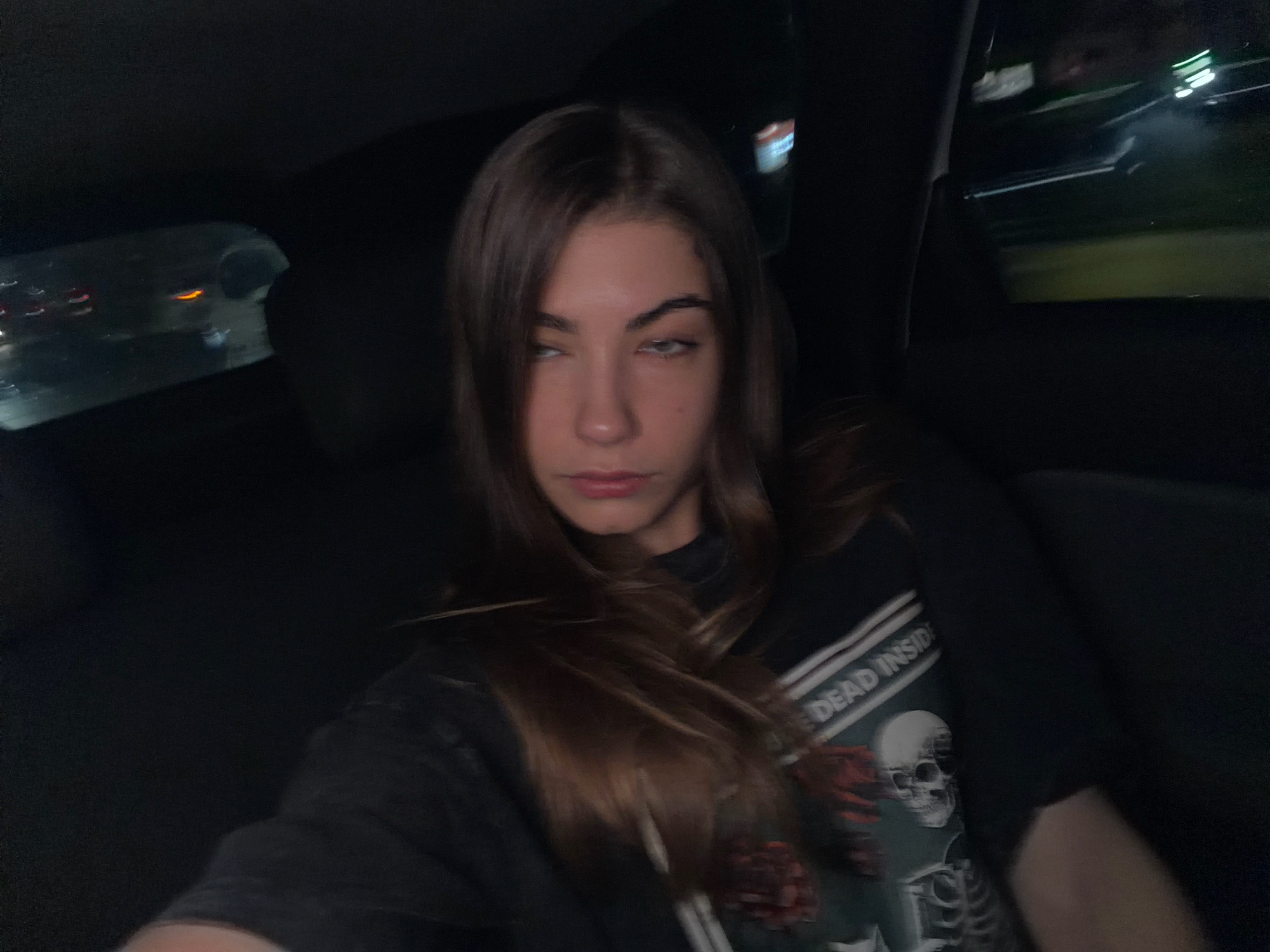 A young woman with long, brown hair is sitting in the back seat of a vehicle at night. She is looking up and to her left with her eyes partially closed. She is wearing a black t-shirt with a graphic design featuring a skeleton and the text "DEAD INSIDE". The interior of the car is dark, with the exception of some faint lights visible through the windows, suggesting they are in motion. The background outside the car appears to be a blurred cityscape with streetlights and building lights. The overall lighting suggests it is nighttime.
