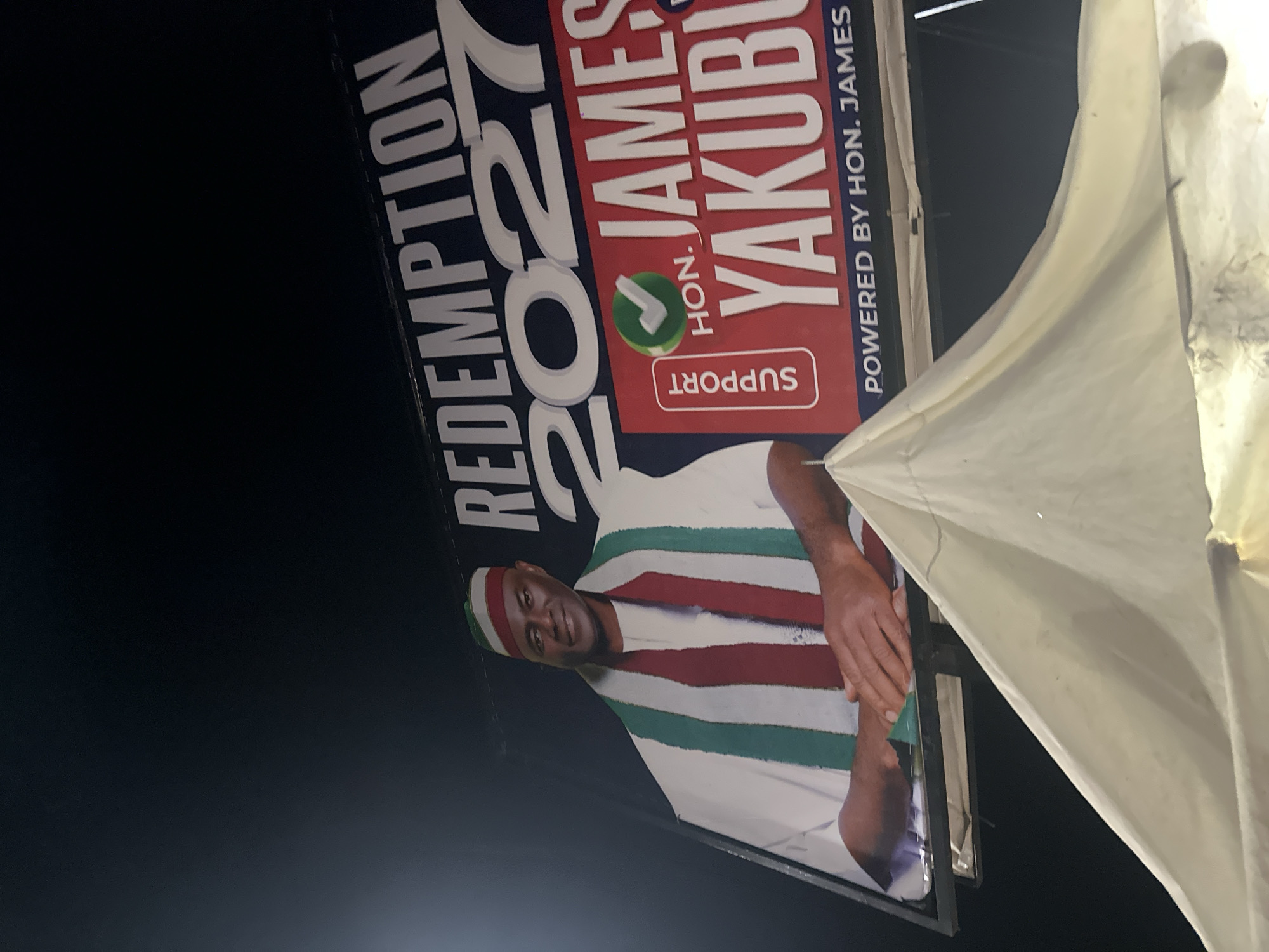 This is a photograph of a political billboard in Gwong, Nigeria. The billboard features a large portrait of a man wearing a traditional cap and a striped robe in white, green, and red. The man is smiling and looking towards the camera. The billboard has large white text that reads "REDEMPTION 2027" and "HON. JAMES YAKUBU". Below the name, there is a green circle with a white checkmark, and a white rectangle with the word "SUPPORT" in red text. At the bottom of the billboard, it says "POWERED BY HON. JAMES". The billboard is partially obscured by a large, cream-colored tent or fabric structure on the right side of the image. The background is dark, suggesting it is nighttime. The overall tone is a political campaign advertisement.