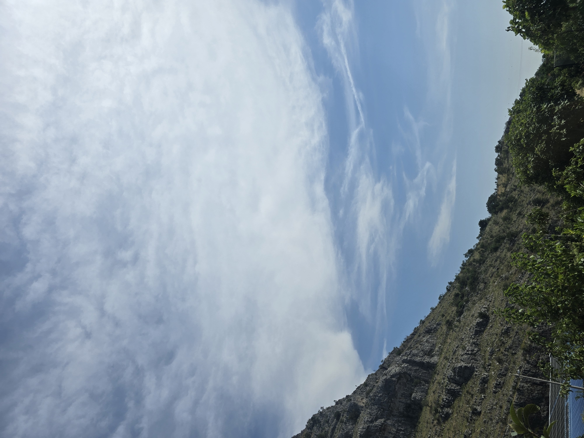 The image captures a natural scene dominated by a vast sky and a rocky mountainside. The sky is a clear blue, streaked with wispy white clouds that create a sense of depth and movement. The sun appears to be high in the sky, suggesting it is daytime, possibly mid-morning or afternoon. The weather seems pleasant, with the clear sky and visible clouds indicating fair conditions.
The mountainside is rugged and covered with sparse vegetation, primarily small trees and bushes clinging to the rocky slopes. The texture of the rock is rough and uneven, showcasing the natural erosion and geological formations. The top edge of the mountainside is lined with denser greenery, suggesting a transition to a more fertile area or a change in elevation.
In the lower right corner of the image, a glimpse of a man-made object is visible. It appears to be a fence or barrier, possibly made of metal and blue netting, hinting at human presence or infrastructure in the vicinity. There are also some leaves from a plant in the foreground, adding a touch of natural detail.
The overall impression is one of a natural landscape, possibly a hiking trail or a viewpoint overlooking a mountainous region. The image evokes a sense of peacefulness and the grandeur of nature, with the vastness of the sky contrasting with the solid, ancient presence of the mountain. There is no visible text in the image.