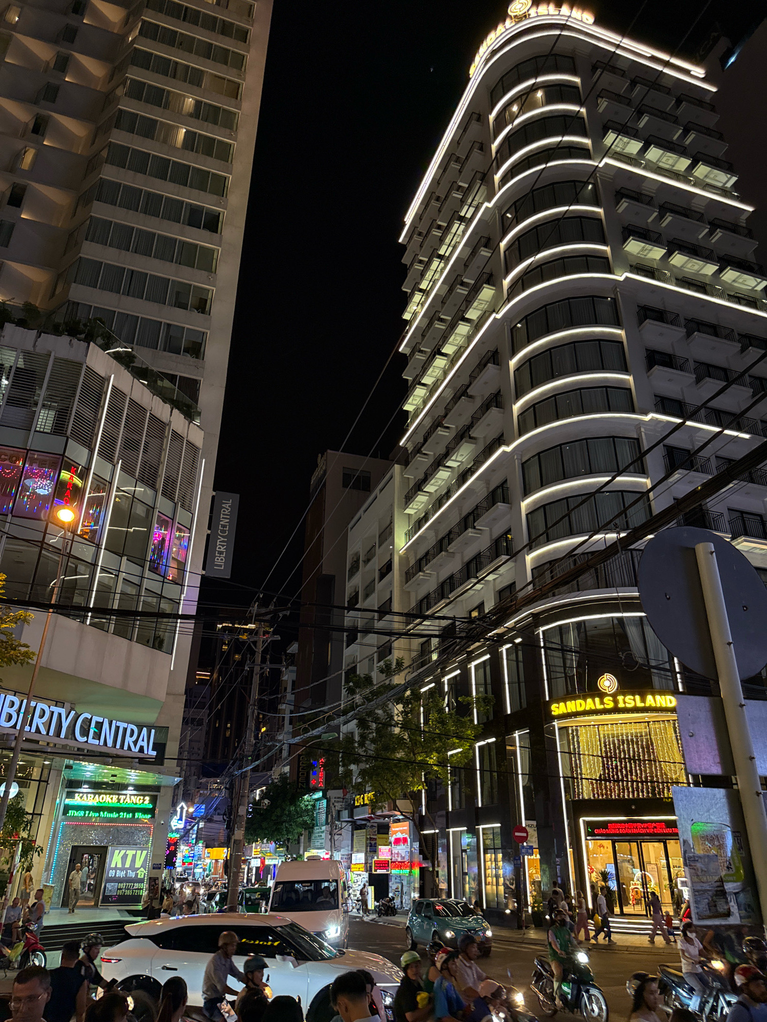 This is a nighttime street-level photograph of Nha Trang, Vietnam, focusing on the intersection of Tran Phu and Hung Vuong streets. The scene is dominated by two large, modern buildings illuminated with white and yellow lights. On the right, the "Sandals Island" building features a curved facade with multiple balconies and extensive decorative lighting. To the left, another building, labeled "LIBERTY CENTRAL," has a more angular design and displays a prominent sign for "KARAOKE TANG 2." The street is busy with traffic, including cars and numerous motorbikes, many with riders wearing helmets. Pedestrians are visible walking on the sidewalks and crossing the street. Various commercial signs and advertisements are displayed on the storefronts, adding to the vibrant urban atmosphere. The sky is dark, indicating late evening or night. The overall impression is one of a bustling, brightly lit commercial and entertainment district.