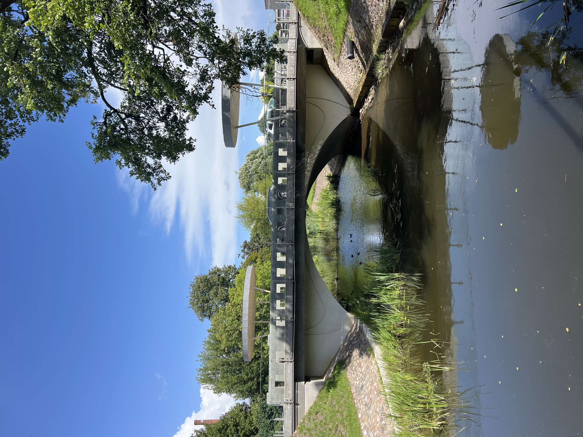 The image depicts a contemporary concrete bridge spanning a calm canal in Saldus, Latvia. The bridge features a sleek, curved design with a wide deck. Along the top of the bridge's railing, there are two distinctive, circular, umbrella-like structures supported by slender poles, suggesting a modern aesthetic. Several vehicles, including what appear to be SUVs, are visible on the bridge deck, indicating it is a functional roadway.
The canal below reflects the sky and the bridge, with a gentle flow. The banks of the canal are lined with green reeds and tall grass on the left, and a stone embankment transitions to grassy slopes on the right. In the water, a few dark-colored ducks are swimming.
The surrounding environment is lush and green, with mature trees visible behind the bridge, creating a natural backdrop. The sky is a clear blue with scattered white clouds, indicating fair weather. On the far left of the image, a tall, red brick chimney can be seen rising above the trees, hinting at industrial or historical structures in the vicinity. The overall scene presents a harmonious blend of modern infrastructure and natural elements within an urban setting.