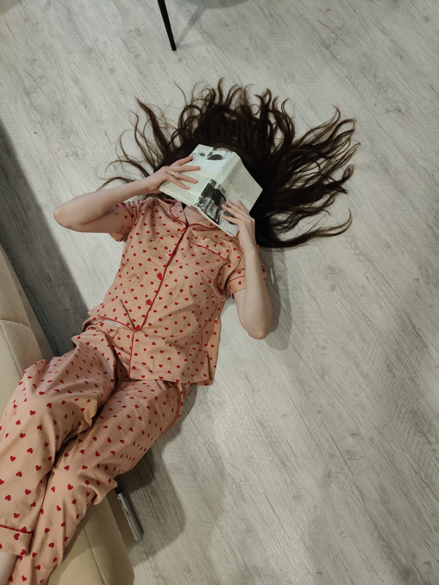 An overhead shot captures a person lying on their back on a light wooden floor, clad in light pink pajamas adorned with small red hearts and red piping. Their long, dark brown hair is spread out around their head. An open book is held over their face, obscuring it entirely. Their arms are bent at the elbows, supporting the book, and a ring is visible on the finger of their left hand.
The setting appears to be an indoor space, likely a home, with a light-colored wooden floor featuring a subtle grain pattern. Part of a beige piece of furniture, possibly a sofa or chair, is visible on the left side of the frame, casting a shadow on the floor. A thin, dark object, potentially a furniture leg or stand, is seen in the upper right corner. The person's posture suggests relaxation or rest, possibly having fallen asleep while reading, or simply enjoying a quiet moment. Text and images are visible on the pages of the book, but they are too small and indistinct to be legible. The lighting suggests it is daytime or early evening indoors.