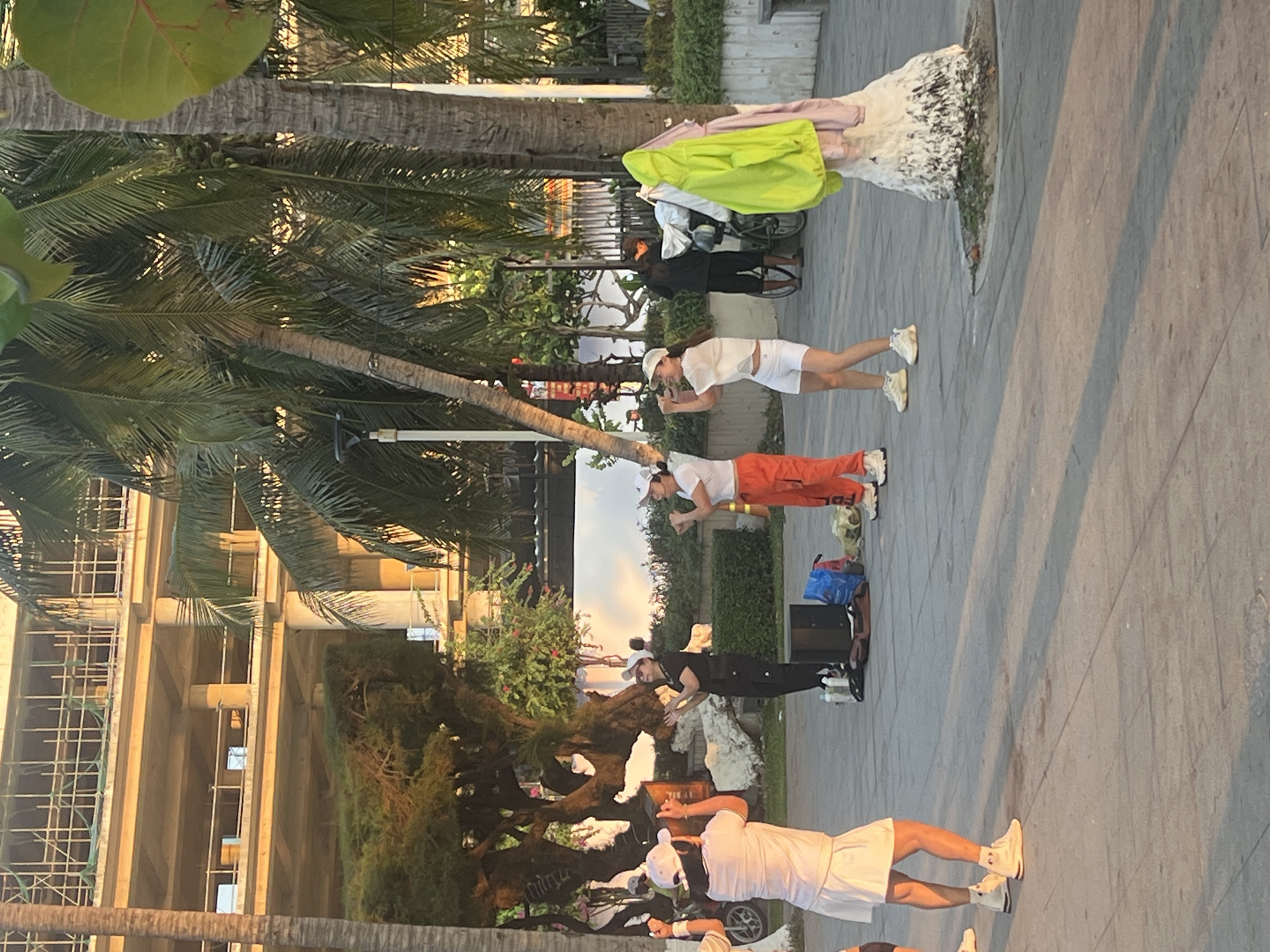 This image captures an outdoor scene, likely a promenade or street in Nha Trang, Vietnam, during the late afternoon or early evening, characterized by warm, soft lighting and long shadows on the paved ground. The setting features numerous palm trees and lush tropical vegetation, with modern buildings, one showing visible scaffolding, in the background.
Several people are present, engaged in what appears to be a street performance or group activity. In the foreground, a person in a white top, white shorts, and a white cap faces away from the camera, standing with bent knees. Closer to the center, two individuals face the camera with their hands near their chests and bent knees; one wears a white top, orange pants (with "FOL" visible on the leg), and a white cap, while the other is dressed in black with a white cap. A black speaker and bags are placed on the ground next to them.
Further back, another person in a white top, white shorts, and a white cap is seen, seemingly adjusting something or observing. To the far right, a person in dark clothing stands next to a bicycle, with brightly colored clothes—a light green jacket, pink fabric, and white items—draped over what appears to be a stand or another bicycle.
The overall atmosphere suggests a lively, outdoor recreational or entertainment event in a tropical urban setting.