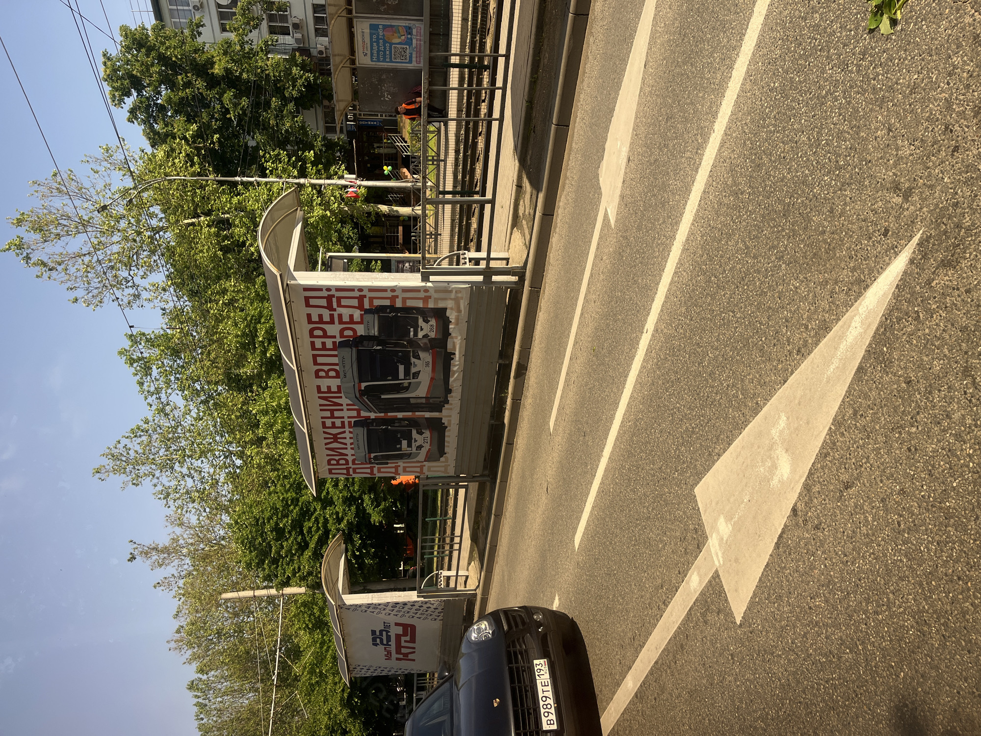 The photograph shows a street scene in Krasnodar, Russia. In the foreground, an asphalt road with a large white arrow indicates a direction of travel. A black car with license plate "B989TE 193" is partially visible. A bus stop with advertisements in Russian, including the phrases "ДВИЖЕНИЕ ВПЕРЕД!" and "МУП-КТТУ", is located alongside the road. The advertisement also features images of modern trams. Behind the bus stop, there are trees and a building. Near the background there is a railroad track. Two people can be seen on the train tracks and on the sidewalk near an orange traffic barrel. The weather appears sunny and clear.