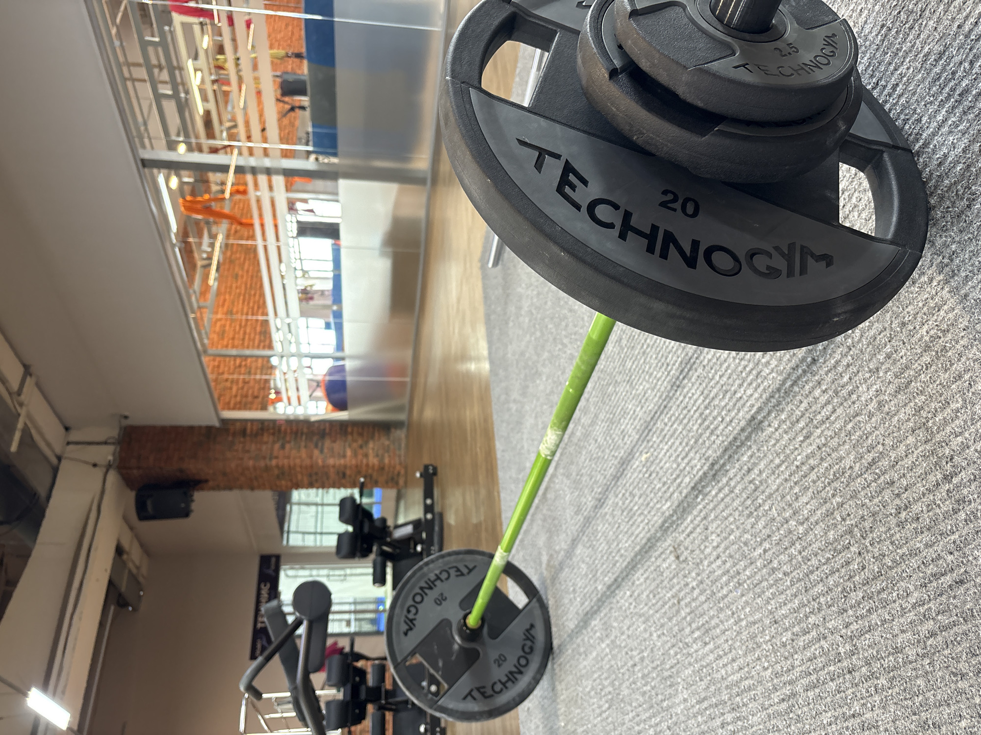 The image shows a barbell with Technogym weight plates in what appears to be a gym. The weight plates have markings indicating 20 kg and 2.5 kg. The barbell has a bright green shaft. The floor is covered with gray carpet tiles. In the background, workout machines, a mirror and an exposed brick wall are visible. Based on the visual information, it is not possible to definitively determine the location in the city of Chaikovsky, Russia.