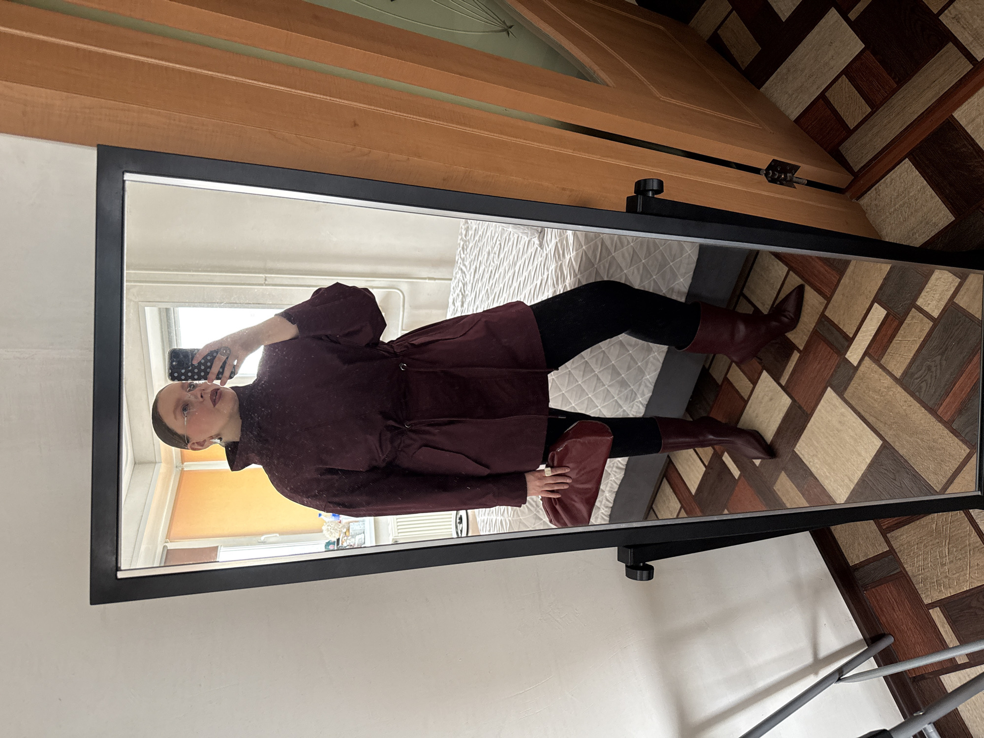 A person is taking a mirror selfie in what appears to be an indoor setting. The person is wearing a burgundy jacket, black leggings, and burgundy boots. They are also holding a matching burgundy purse. The mirror reflects a portion of a room with a bed covered in a white quilted comforter, a window, and what appears to be a tiled floor. The wooden door behind the mirror suggests an interior space. There is no information in the photo to verify its location as Izhevsk, Russia.