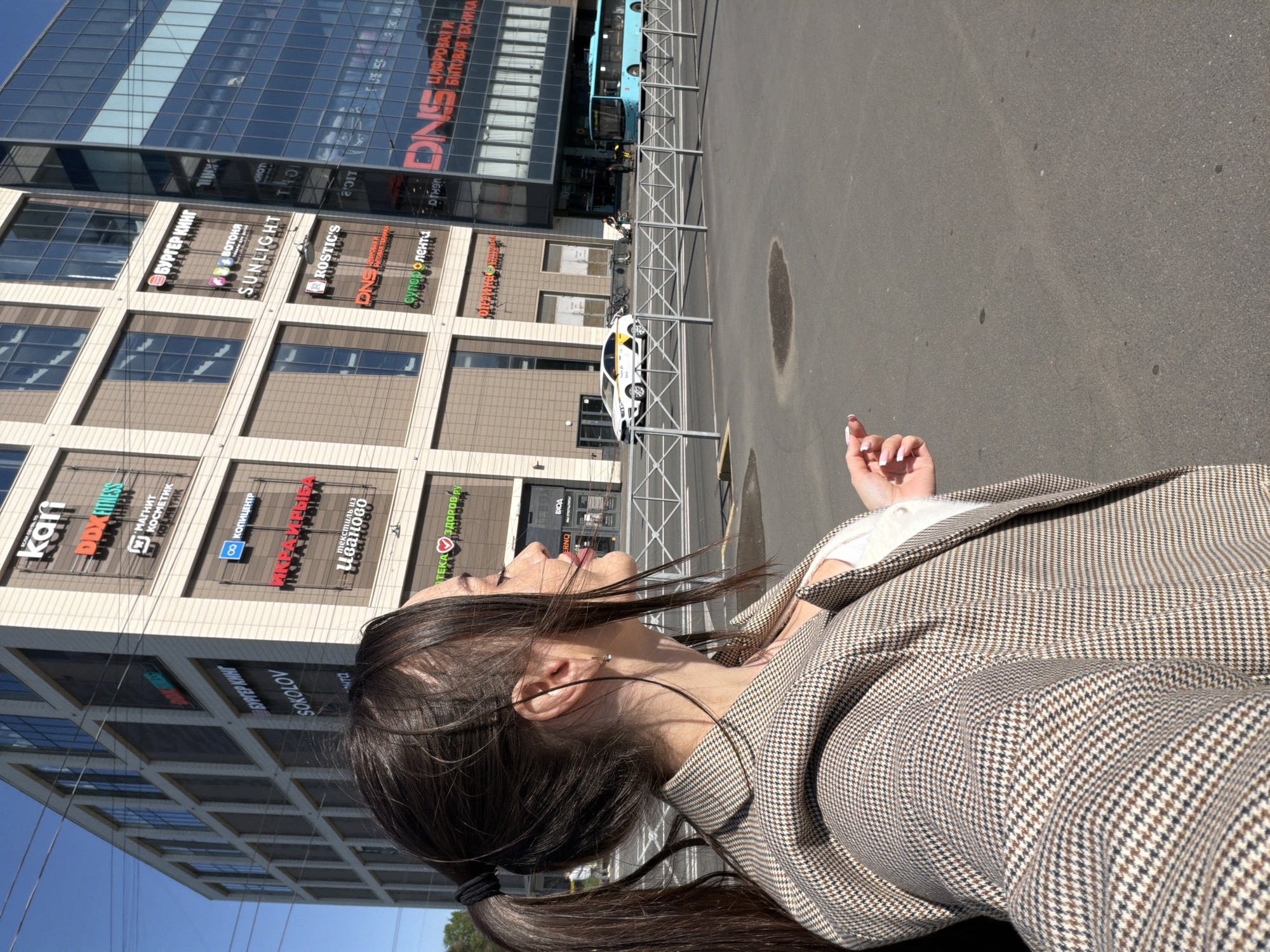 A woman stands on a city street in Saint Petersburg, Russia. She wears a brown and white patterned jacket. Her dark hair is pulled back into a ponytail. The building behind her contains various storefronts, including Kari, Burger King, SUNLIGHT, Rostic's, and DNS. A white taxi cab is parked on the street, and a bus is visible further down. The blue sky indicates it is daytime.