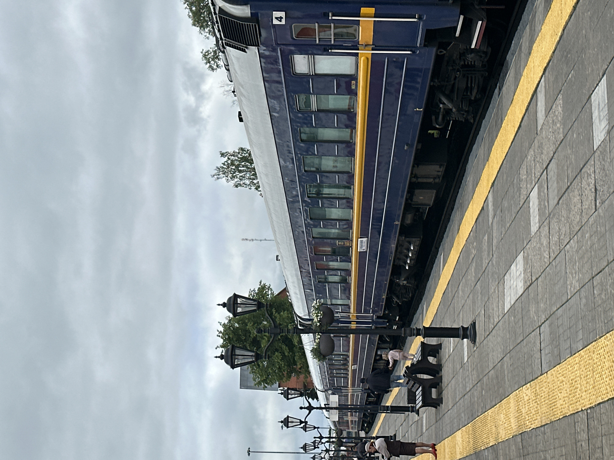 This is a photograph taken at the Sortavala railway station in Sortavala, Russia. The image features a dark blue train with a yellow stripe along its length, stopped at the platform. The platform is paved with gray tiles and has a prominent yellow tactile warning strip running along the edge. Several vintage-style lampposts with ornate designs are positioned along the platform, with some holding hanging flower baskets.
In the foreground, a woman wearing a light-colored hat and a brown skirt suit stands on the platform. Further down the platform, a man and a young child in light pink clothing are walking towards the train. A dark-colored bench is visible on the platform. In the background, trees and a building with a red roof are present. The sky is overcast, suggesting a cloudy day. The train car visible has the number "4" prominently displayed. A sign on the train car reads "MECT90". The overall scene depicts a quiet moment at a railway station with passengers preparing to board or disembark.