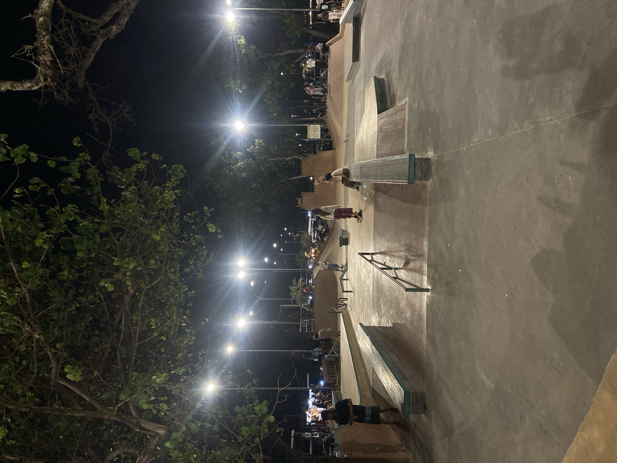 The image depicts a skate park in Kuta, Indonesia, at night. The area is well-lit by streetlights. Several people are scattered throughout the park, with some appearing to be skateboarding or observing. The skate park features various ramps, rails, and concrete surfaces. In the periphery, trees frame the scene, adding a natural element. There are some parked cars in the distance.