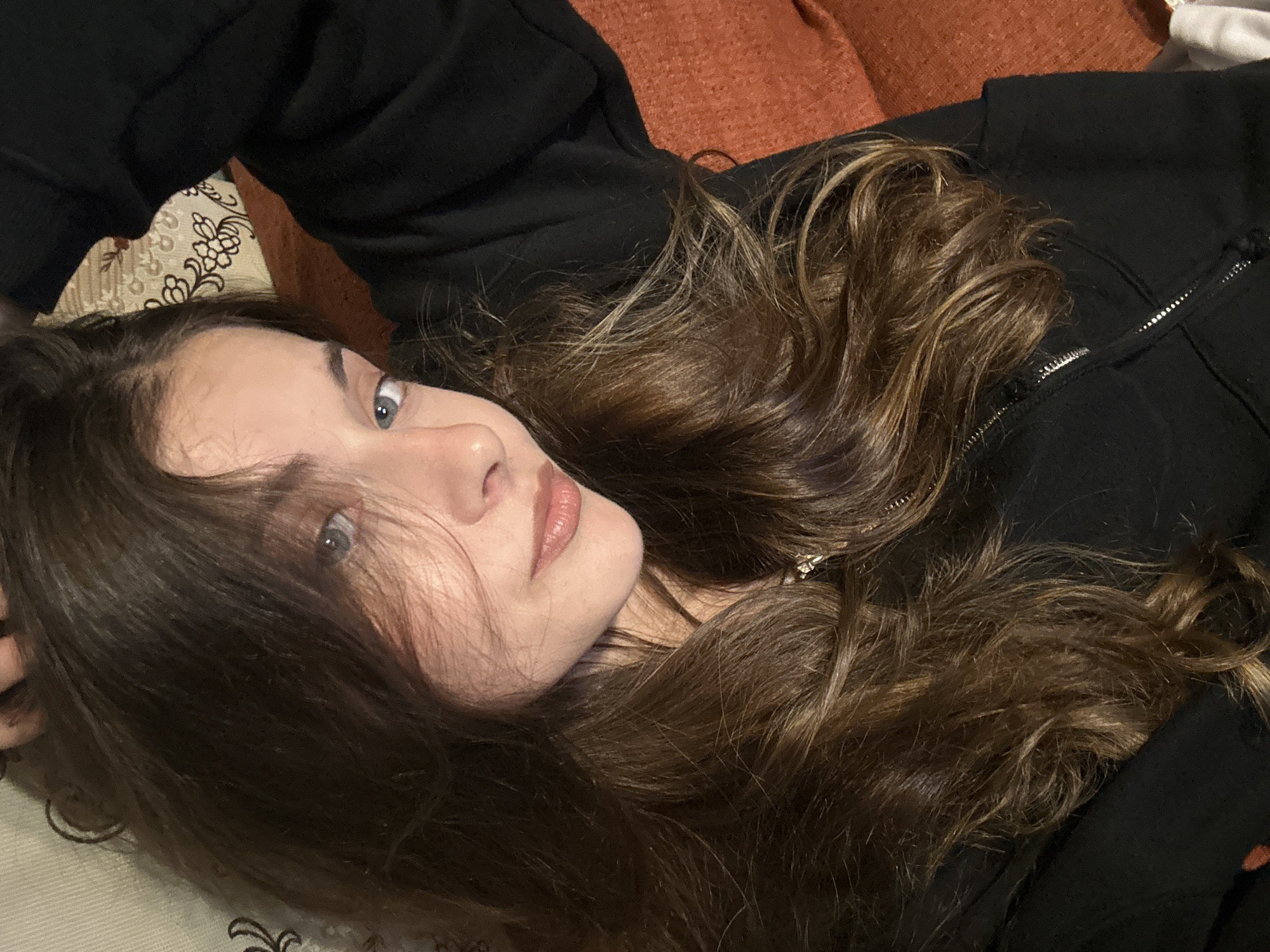 A young female with long, wavy brown hair and lighter highlights, and blue eyes, is depicted in a close-up, diagonal shot. She is reclining, with her head positioned towards the upper left of the frame and her body extending towards the lower right. Her gaze is directed at the camera. Her right hand (viewer's left) is raised, with fingers partially visible near the top of her head amidst her hair. She is wearing a black zippered hoodie or jacket, with the zipper and fabric texture visible on the lower front. Her lips have a natural or light nude-pink color. The background consists of a light-colored patterned fabric, possibly a pillow with a brown floral design, in the upper right quadrant, and a textured, rust-orange fabric, likely a sofa or blanket, in the lower right. The scene is located in Unknown, Unknown.