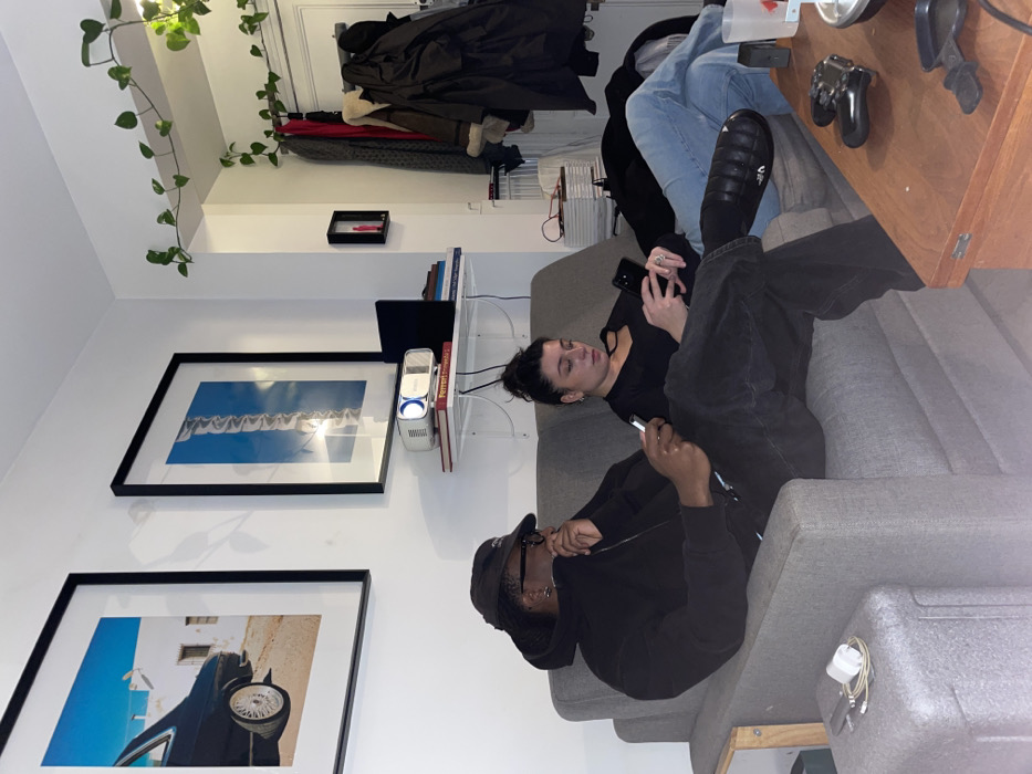 Two people are lounging on a gray couch, both engrossed in their phones. One is a woman in a black top and the other is a man wearing a black hoodie and cap. Next to the couch is a wooden table with a game controller and other items. On the wall behind the couch hang two framed pictures and a shelf with books and a projector. A leafy vine drapes from the ceiling, and a coat rack stands near the top of the frame.