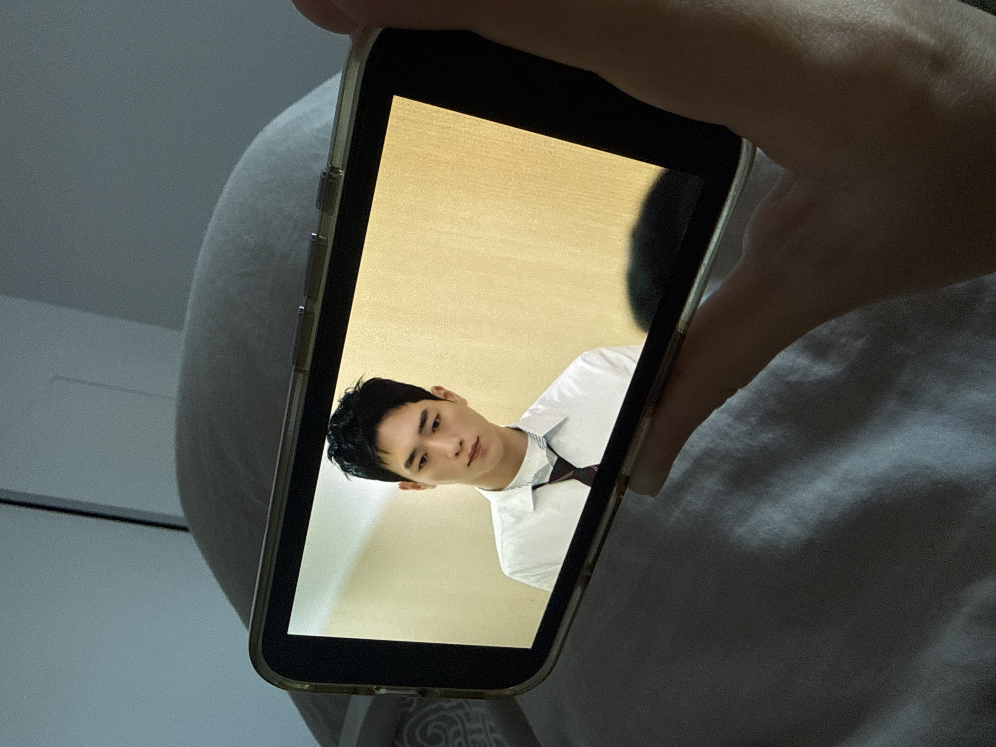 A person is holding a smartphone displaying an image of a young man in a white shirt and tie. The man on the screen has dark hair and is looking towards the camera. The background on the phone screen is a light beige color. The phone is being held over what appears to be a white or light-colored cushion or blanket. The environment appears to be indoors. There are no visible landmarks indicating the location is Krasnoyarsk, Russia.