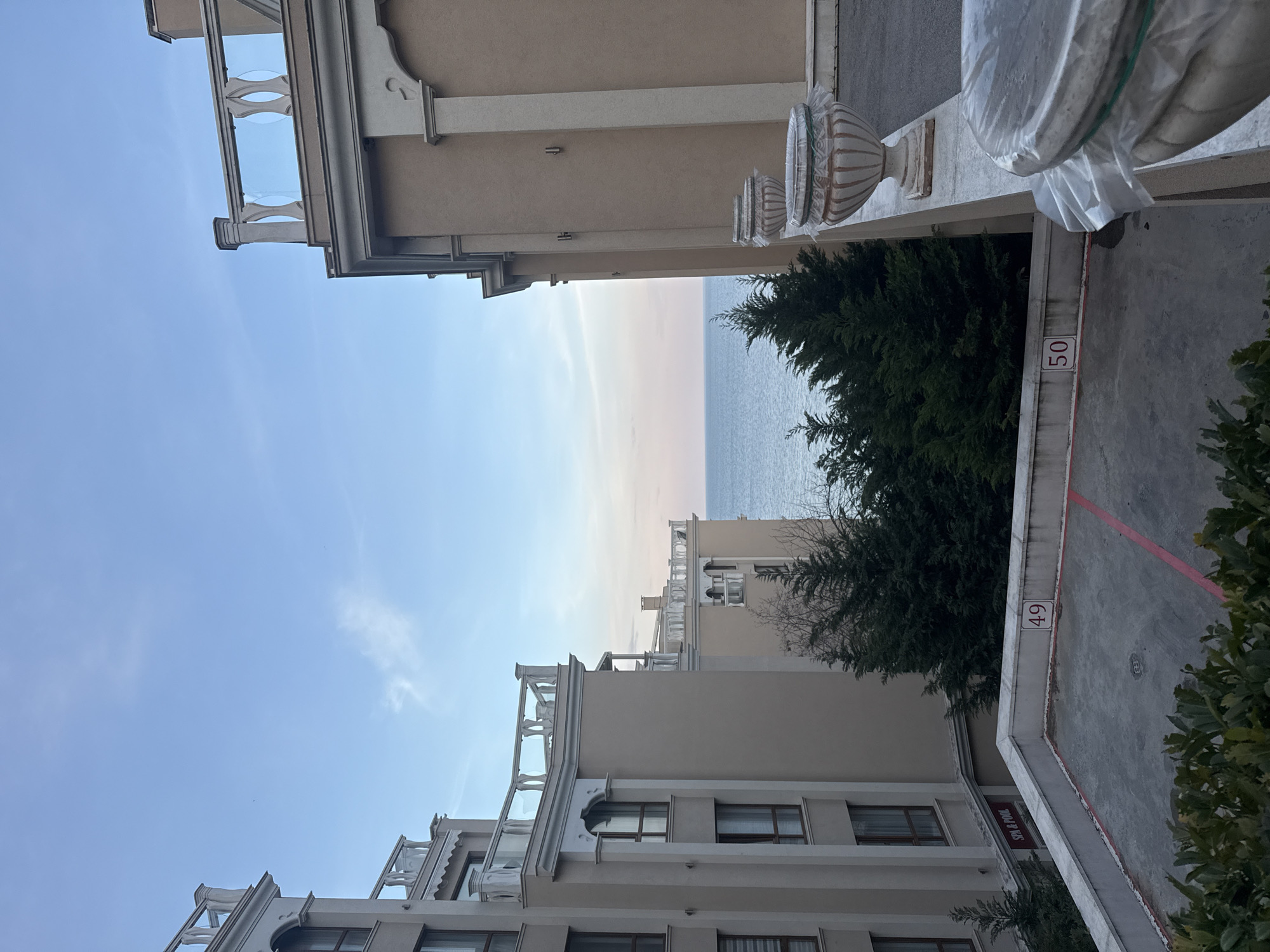 The image presents a view between two multi-story buildings in Varna, Bulgaria. The structures are constructed of tan-colored stucco. The left and lower portions of the image shows architecture with white-trimmed windows, balconies with decorative balustrades, and projecting cornices. The right and upper part of the image has a similar building profile, with decorative urns and a parking area marked with numbers '49' and '50'. In the gap between the buildings, a strip of the Black Sea and sky are visible. Green trees are growing between the buildings.