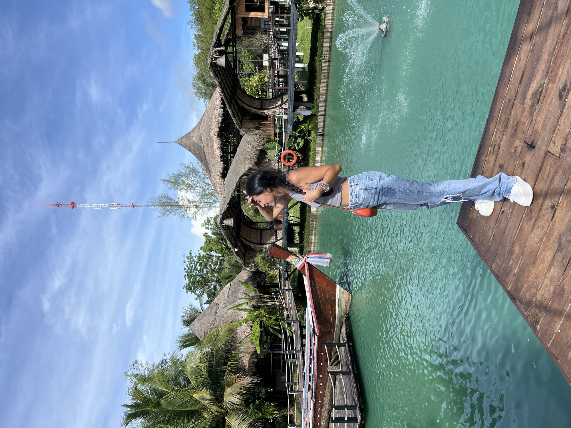 A young woman stands on a wooden pier next to a body of turquoise water. She is wearing ripped blue jeans and a gray tank top, with a small orange purse slung over her shoulder. Her hair is dark and falls to her shoulders, and she appears to be shielding her eyes from the sun, suggesting a bright, sunny day. The water features a fountain in the distance.
Behind the woman are several structures with thatched roofs, reminiscent of traditional Southeast Asian architecture. Lush green palm trees and other tropical foliage surround the buildings. A long, wooden boat with colorful trim is docked nearby, tied to the pier. Further in the background, a tall, red and white communication tower rises against a bright blue sky with scattered white clouds.
The scene appears to be a tourist destination, possibly a resort or a recreational area, given the water features, boat, and unique architecture. The time of day is clearly daytime, indicated by the bright sunlight and clear sky. The overall atmosphere seems relaxed and picturesque, capturing a moment of leisure in a tropical setting. The location cues point to Tambon Pa Kae Bo Hin, Thailand.