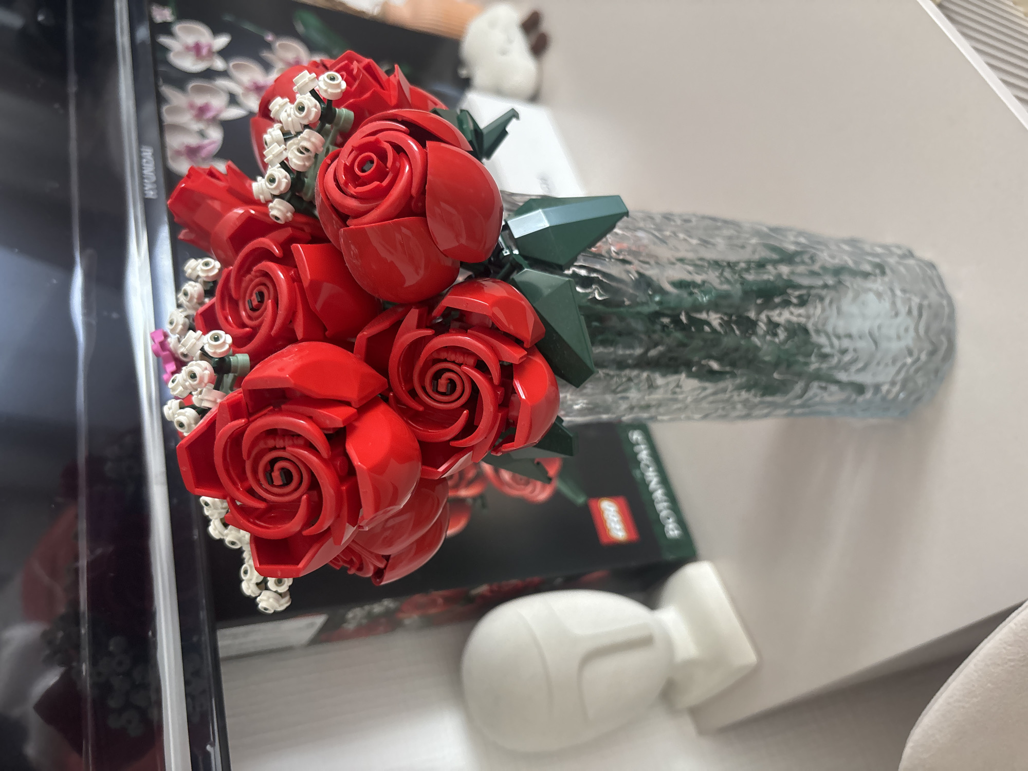 A close-up shot shows a Lego rose bouquet in a textured glass vase, lying on top of a Lego botanicals box. A white, abstract, bulbous object is also on the table next to the box. The scene is set on a white table next to a black object and a white seat. The location is Unknown, Unknown.