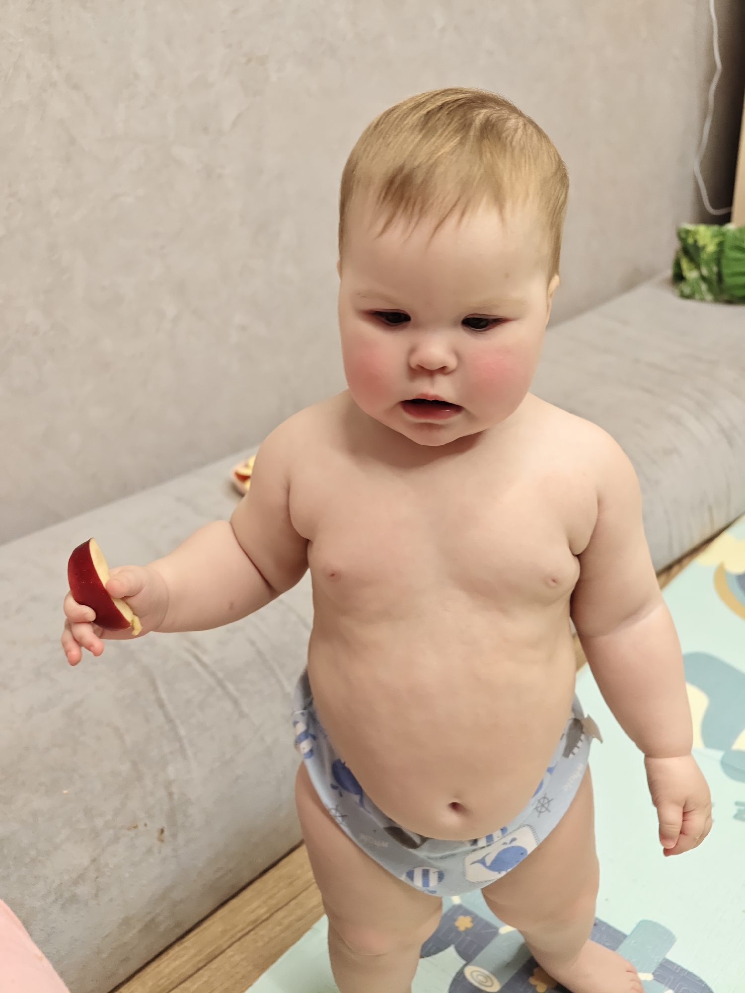 A close-up shot shows a toddler standing indoors. The child has fair skin, blonde hair, and chubby cheeks with a slight flush. The toddler is only wearing a diaper patterned with blue and white marine life. The child holds a slice of red apple in their left hand, looking down at it. The background includes a textured light gray couch or wall, a wooden floor, and a play mat with a blue and gray pattern. The image appears to have been taken at eye level, focusing on the child's expression and gesture. The location is Chaikovsky, Russia.