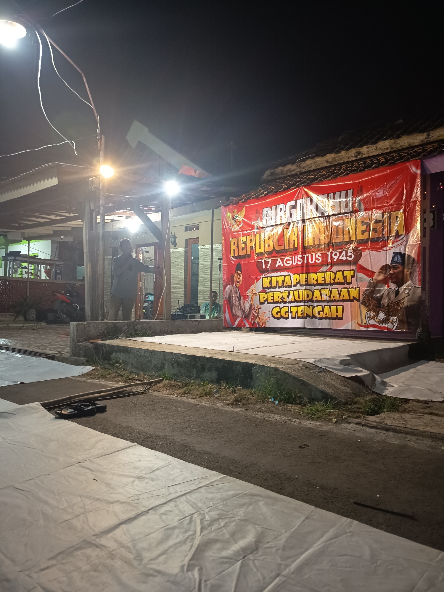 The image captures an outdoor scene at night, illuminated by artificial lights. A large red banner with white and yellow text is prominently displayed, likely for an event. The banner features images of two men in military-style uniforms, one of whom is saluting. Text on the banner includes "DIRGAHAYU REPUBLIK INDONESIA 17 AGUSTUS 1945 KITAPERERAT PERSAUDARAAN GG TENGAH". In the background, a building with a tiled roof is visible. To the left of the banner, a man in a gray shirt and brown pants stands near some wooden structures, speaking into a microphone. A motorcycle is parked nearby. In the foreground, a white tarp covers a surface, and a pair of flip-flops rests on the ground next to some wooden sticks. The ground is a mix of asphalt and concrete. Another person in a green shirt is partially visible in the background on the right. The overall impression is that of a public gathering or celebration taking place at an outdoor venue in the evening.
