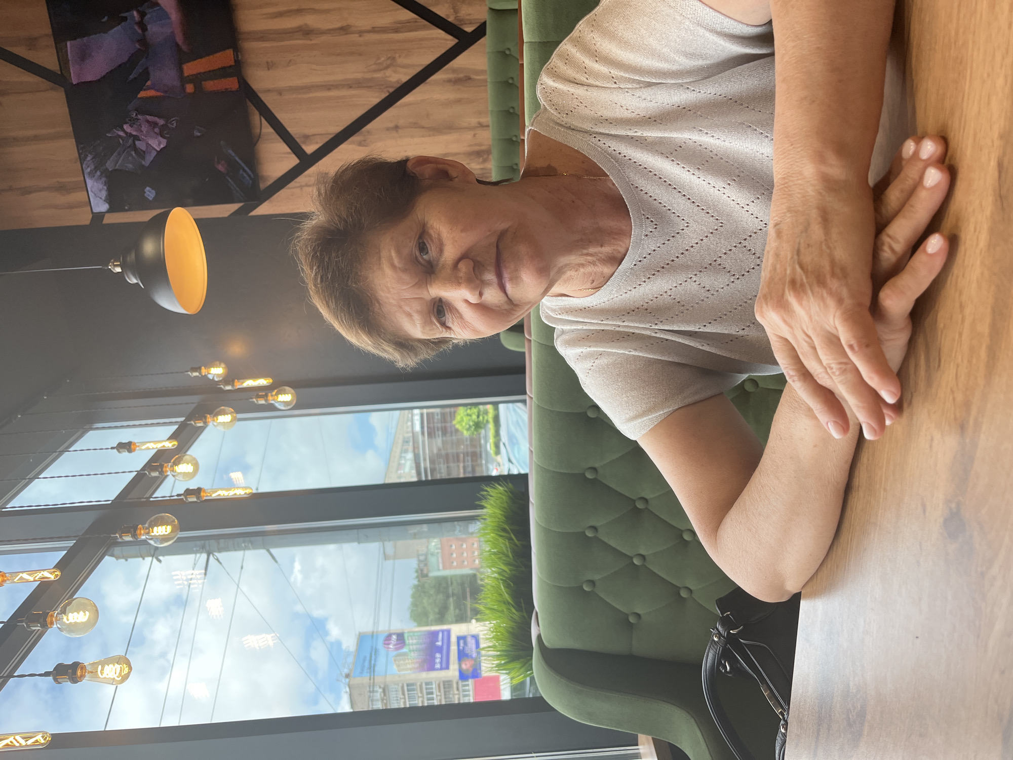 This is a portrait-oriented photograph of an elderly woman sitting at a table in a cafe. The woman is wearing a light-colored, shimmering top and has her hands clasped on the wooden table in front of her. Her expression is neutral, and she is looking towards the camera. Behind her, a green, tufted sofa is visible. The setting appears to be a modern cafe with large windows overlooking a street. Various incandescent light bulbs hang from the ceiling, some with visible filaments, creating a warm ambiance. Through the windows, buildings and trees can be seen, suggesting it is daytime. The sky appears partly cloudy. On the table, a black handbag is partially visible. There is some text on a poster visible through the window, but it is too blurry to read. The location context provided indicates Izhevsk, Russia.
