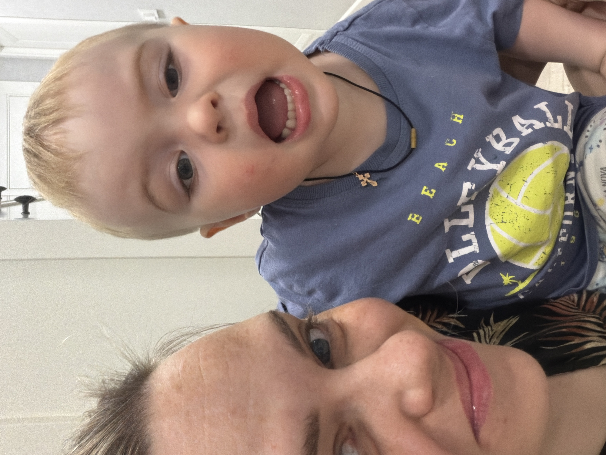 A close-up selfie shows a woman with short, light-brown hair, fair skin, and blue eyes, with a small child in her arms. The child, who has blond hair and fair skin, is wearing a blue T-shirt with a print of a volleyball and the word "BEACH VOLLEYBALL" on it. The child has their mouth open, as if shouting or surprised. The woman is smiling. The background appears to be a plain white wall. No specific details indicate the location is Votkinsk, Russia.