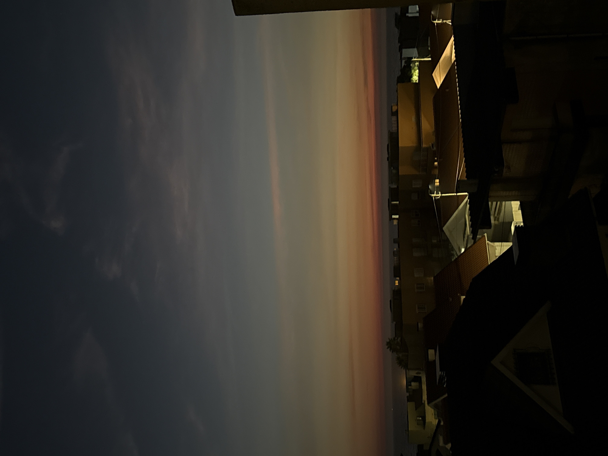 This is a vertical, eye-level shot of a cityscape at dusk, likely taken from a high vantage point. The sky is a gradient of dark blue at the top, transitioning to lighter blues and then to hues of orange and red near the horizon. Faint, elongated clouds are visible in the upper part of the sky.
Below the sky, a body of water, presumably the Atlantic Ocean, is visible in the distance. Along the coastline, a strip of land with buildings is situated. The buildings are a mix of styles and colors. Some are dark and silhouetted, while others are illuminated with warm yellow and white lights, suggesting they are apartments or hotels.
In the foreground, the roofs of several houses dominate the lower half of the image. These structures are dark and appear to be residential. The details of these foreground buildings are obscured by shadow, with only the outlines of their roofs and some architectural features like chimneys and gables being discernible. The overall impression is one of a coastal city settling into evening, with the warm glow of lights contrasting with the deepening twilight sky. The image does not contain any people or discernible actions.