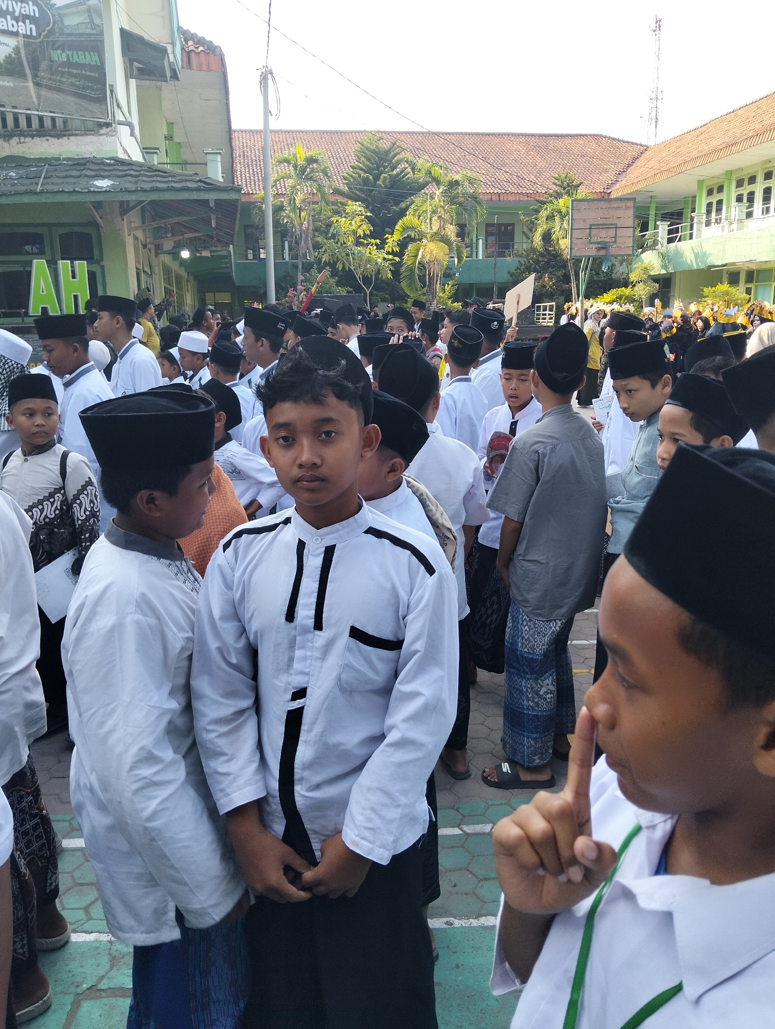 The image captures a gathering of young males, likely students, in what appears to be a schoolyard in Kranji, Indonesia. They are predominantly dressed in white shirts and black peci hats, common attire for Islamic events. The background reveals school buildings with green accents and tiled roofs, suggesting an educational institution. Palm trees and other greenery are visible, indicating a tropical climate. The overall atmosphere seems to be one of a communal event, possibly a religious or educational observance, given the attire and the presence of a large group of similarly dressed individuals. The focus is on the young boys, with some in the foreground looking directly at the camera. The lighting suggests it is daytime, with sunlight casting shadows.