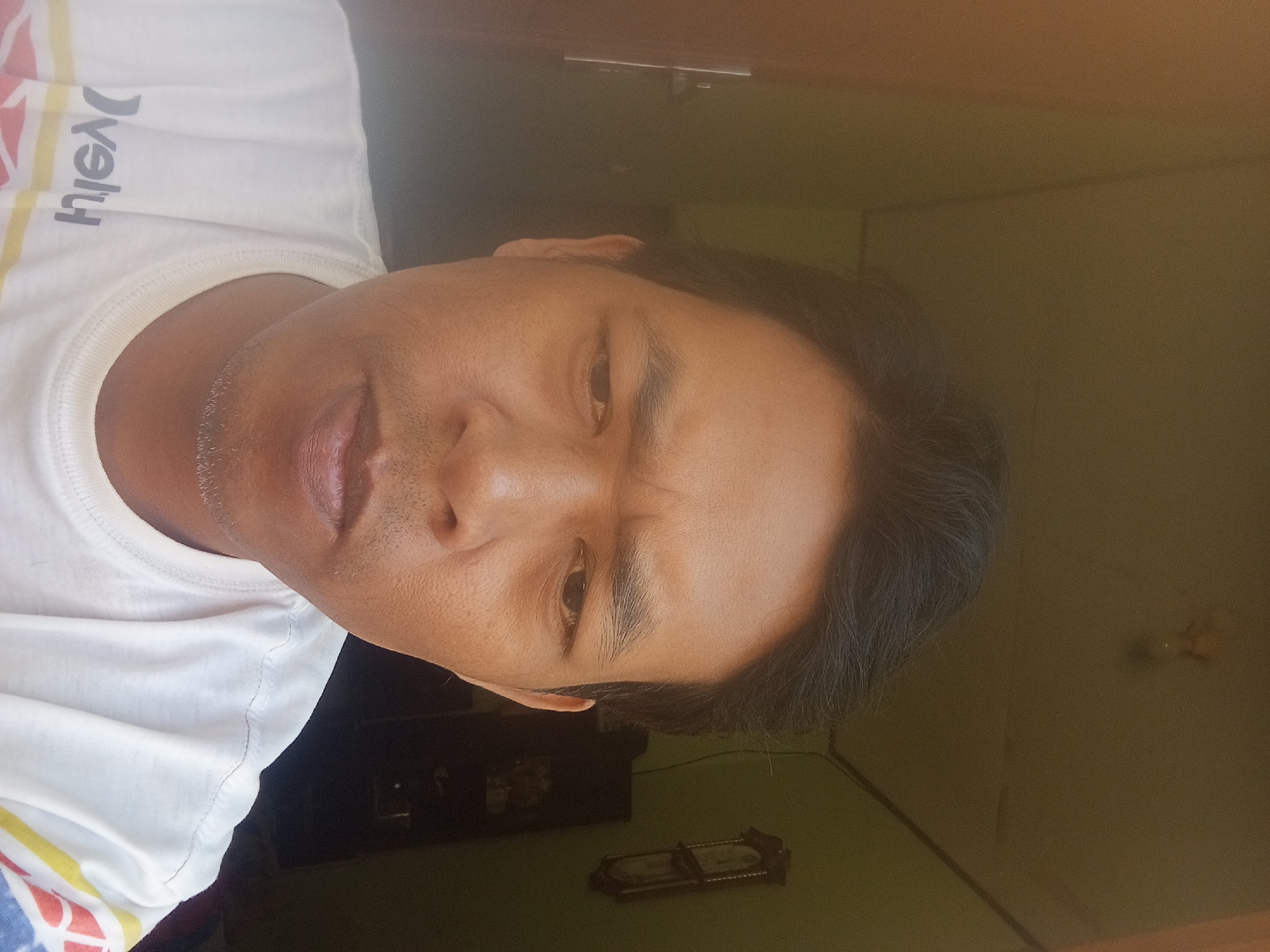 A close-up, waist-up selfie of a man taken indoors. The man has dark hair and fair skin. He is wearing a white t-shirt with a logo on the left side of his chest. The background is a room with a green wall, a dark closet, and a framed clock on the wall. The man is looking directly at the camera with a neutral expression. The image appears to have been taken during daylight hours. The location is likely Pagar Alam, Indonesia, given the user's request.