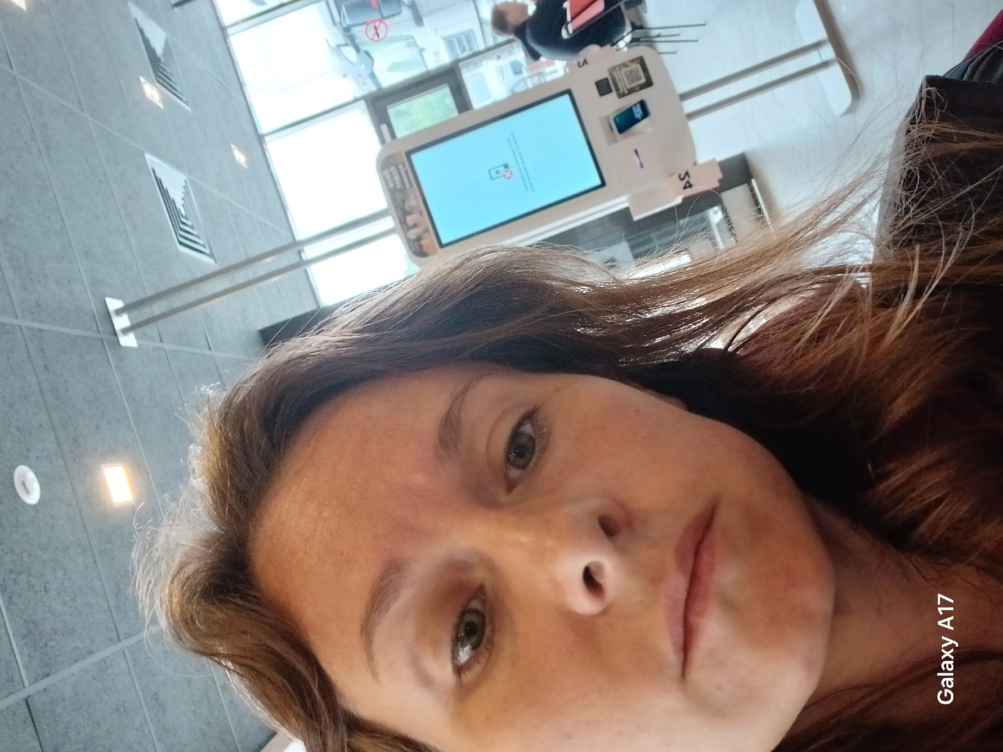 A selfie of a woman in front of a self-service kiosk. The kiosk screen shows an image of a smartphone. The woman is facing the camera. In the background, there are windows and a person sitting at a table. The scene appears to be inside a commercial space. The kiosk has the letters "S4" on the side. It is unclear if the location is Yekaterinburg.