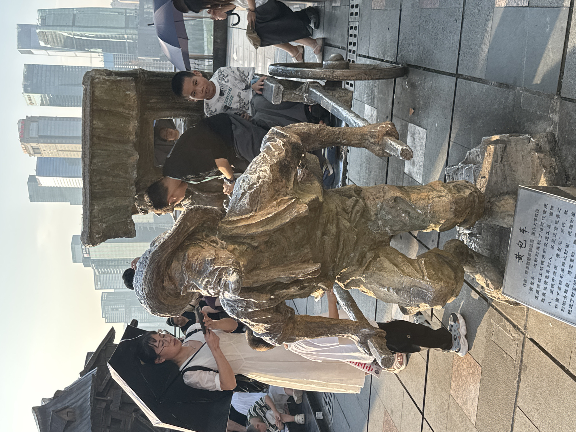 The image captures an outdoor scene in Yuzhong District, China, featuring a bronze sculpture of a person pushing a cart, likely a street vendor or laborer from a past era, set against a backdrop of modern skyscrapers. Several people are present, observing the sculpture and the surrounding urban environment.
In the foreground, the bronze sculpture is the primary subject. It depicts a weathered figure with a hat and what appears to be a beard, leaning forward to push a two-wheeled cart. The cart has a rough, textured surface, and one wheel is clearly visible. The sculpture is placed on a paved ground made of square tiles.
To the left of the sculpture, a woman in a white dress holds an open black umbrella, shielding herself from the sun, and is looking at her phone. A child in a striped shirt is seated on the ground nearby. Behind the woman, another individual is partially visible.
In the middle ground, several people are positioned around the sculpture and the cart. A young boy wearing a patterned t-shirt sits on the cart, looking towards the camera. Behind him, a person in a dark shirt is seated. Further back, other individuals are standing or sitting, some holding umbrellas.
The background is dominated by a dense cluster of modern skyscrapers, indicative of a major urban center. The sky is bright, suggesting it is daytime, possibly late afternoon due to the warm lighting. The overall impression is a juxtaposition of historical representation with contemporary urban life. A plaque with Chinese characters is visible at the base of the sculpture, providing information about the artwork.